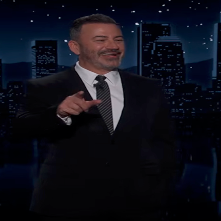 A person in a suit is speaking on a night-time talk show set with a city skyline backdrop