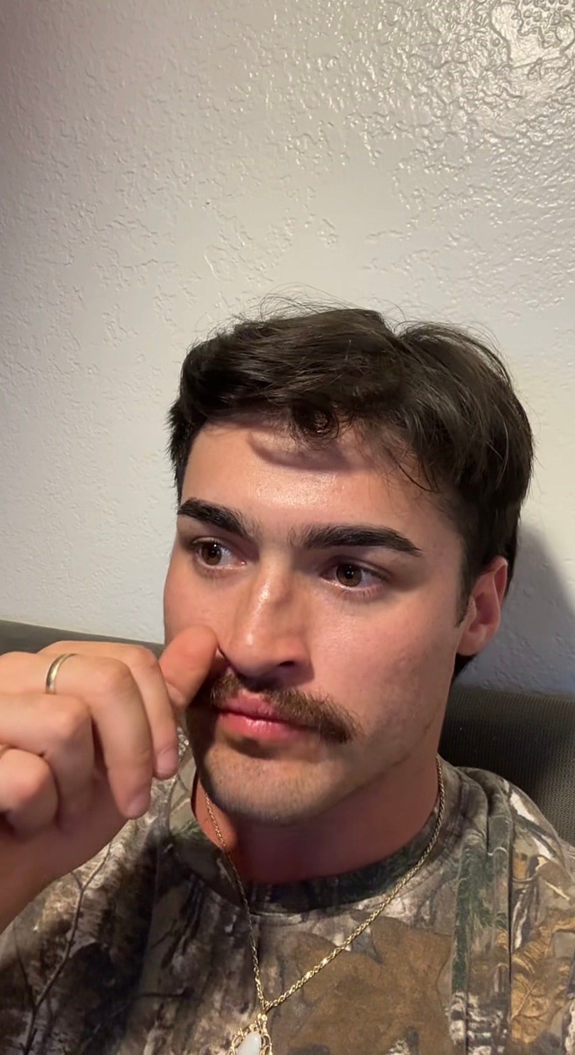 Person with a mustache twirling it with a thoughtful expression, wearing a camouflage-patterned shirt, seated indoors