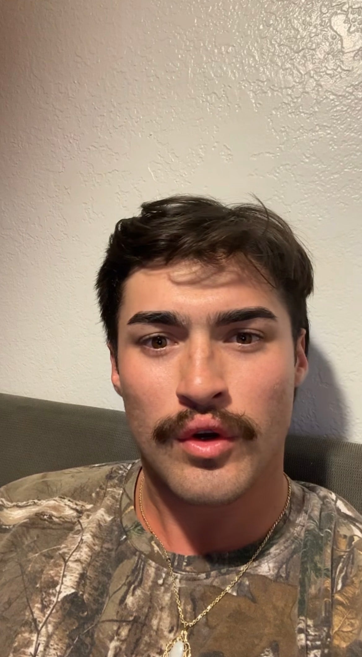 Person with a mustache wearing a camouflage-patterned shirt and a gold chain, sitting on a couch