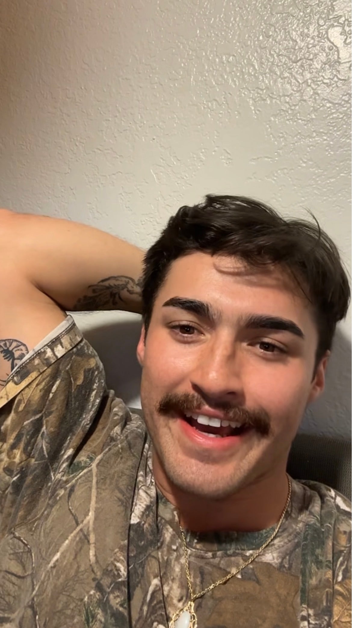 A person with a mustache and tattoos relaxes, smiling, wearing a camo-patterned shirt and a gold chain necklace