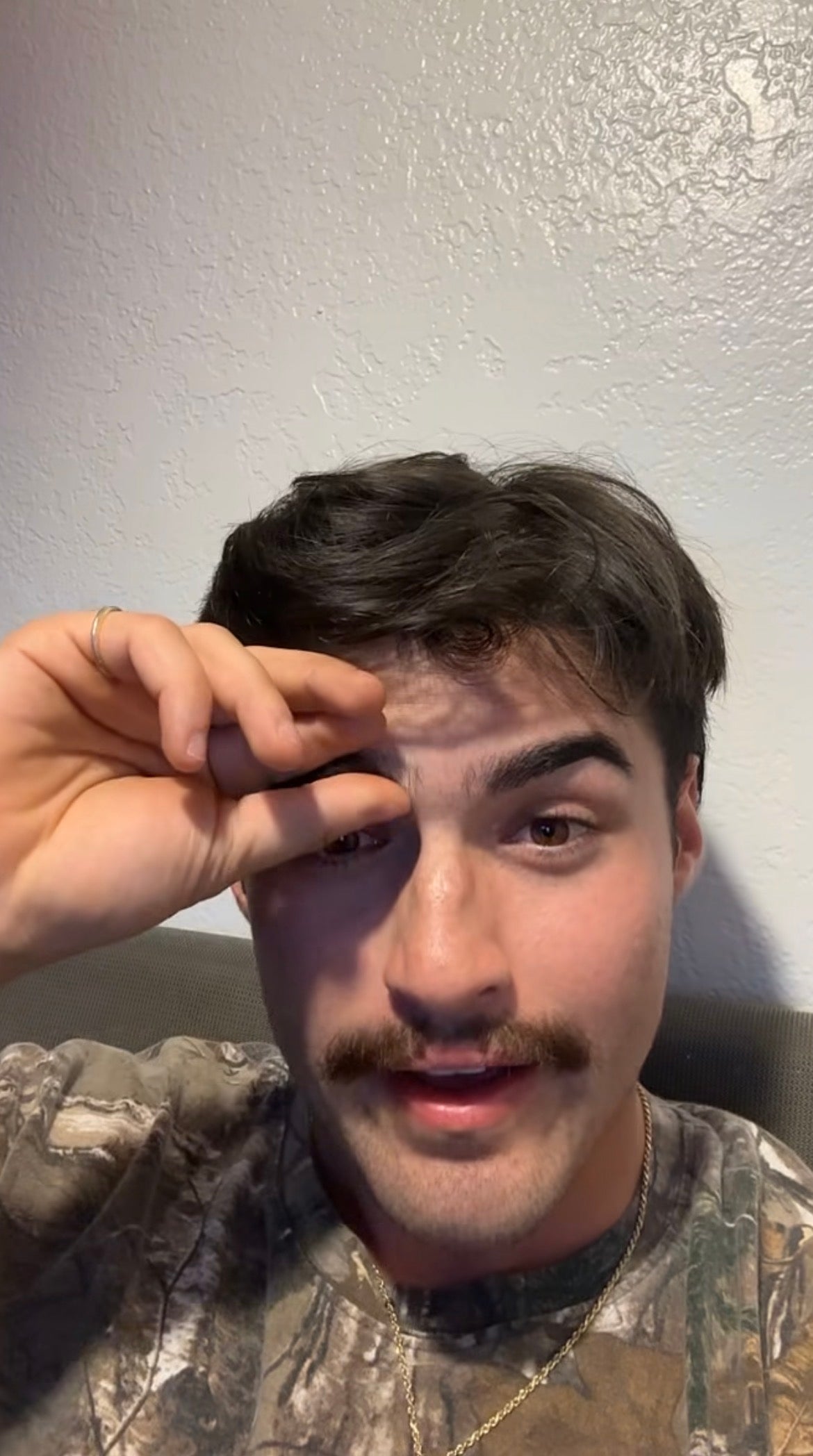 A person with a mustache is touching their eyebrow, wearing a camouflage-patterned shirt, sitting against a plain wall
