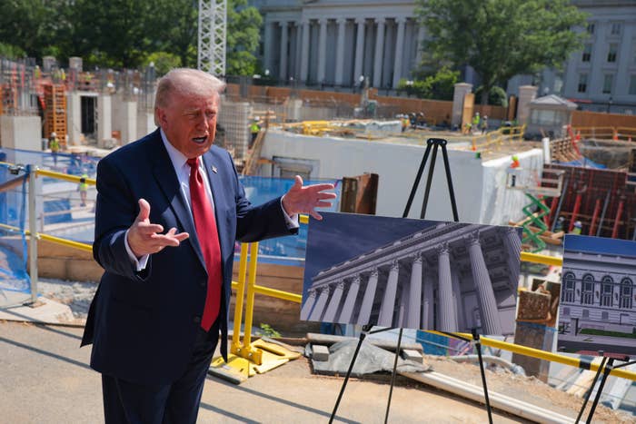 A man in a suit gestures near construction site displays with architectural photos, speaking energetically outdoors