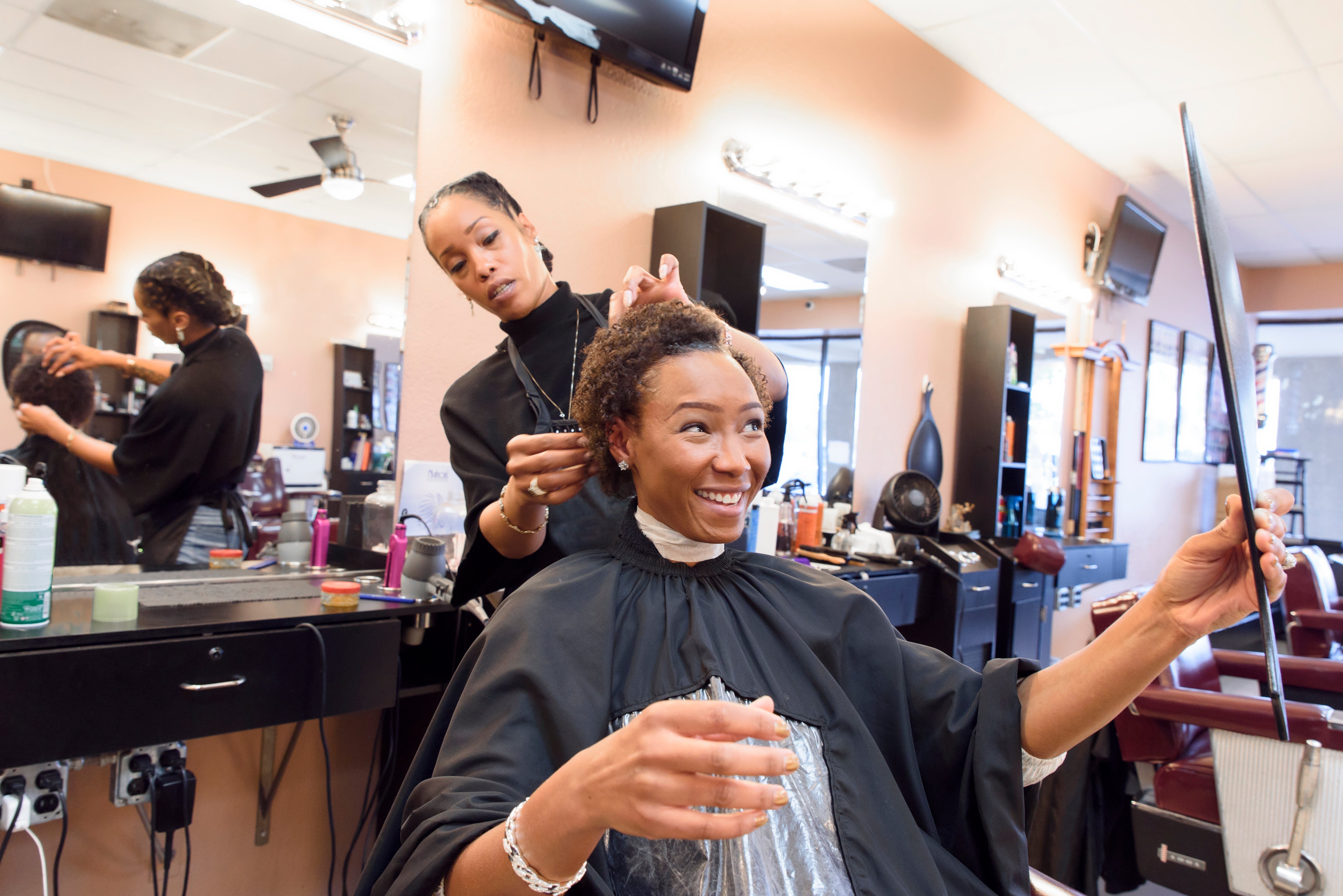 A person smiles while having their hair styled at a salon, holding a mirror to check the progress. Stylist is focused on their work