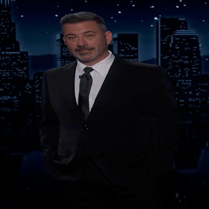 A person in a suit stands on a stage with a cityscape backdrop, likely during a talk show