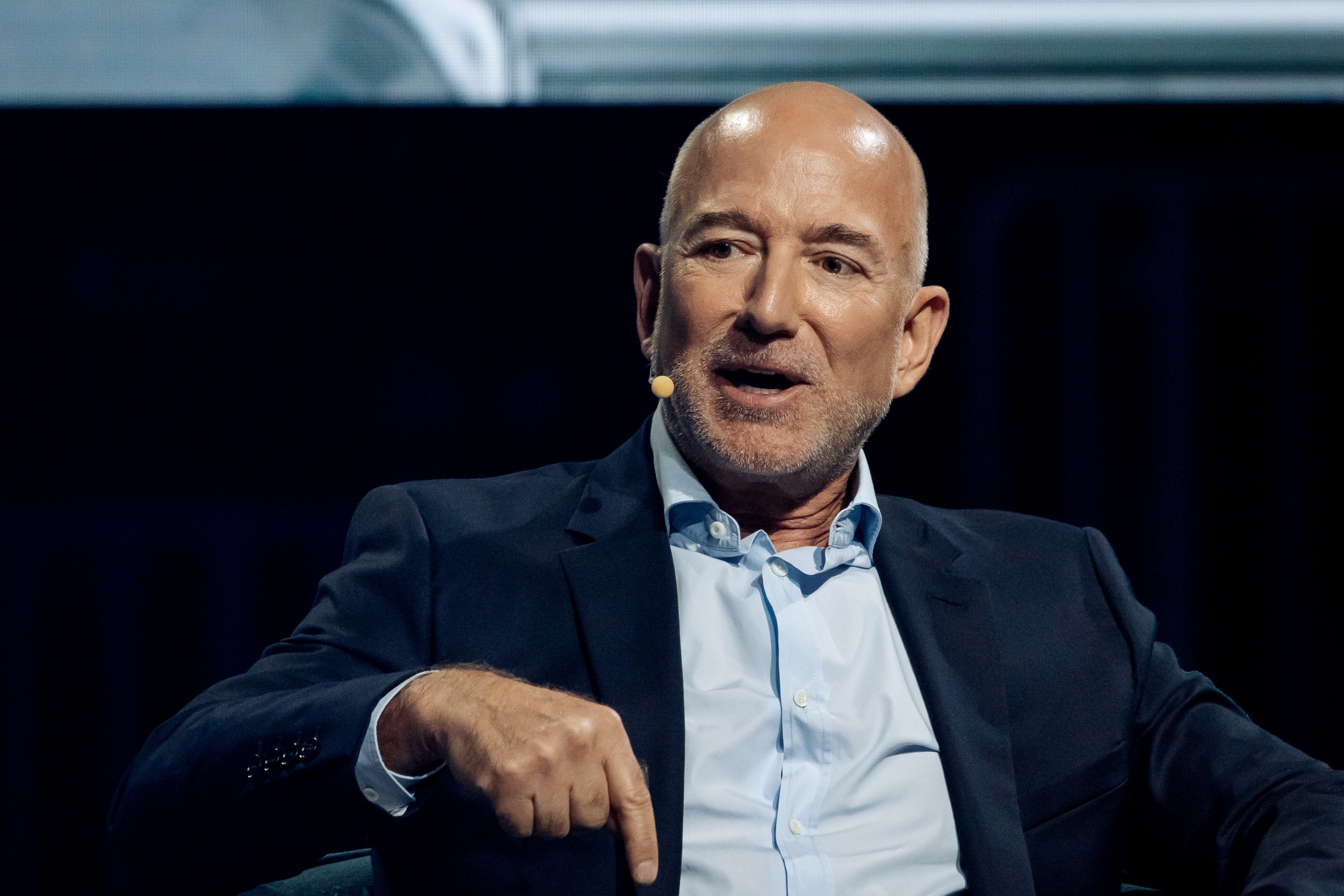 A bald man in a suit speaks on stage with a microphone headset, gesturing with his hand during a panel discussion