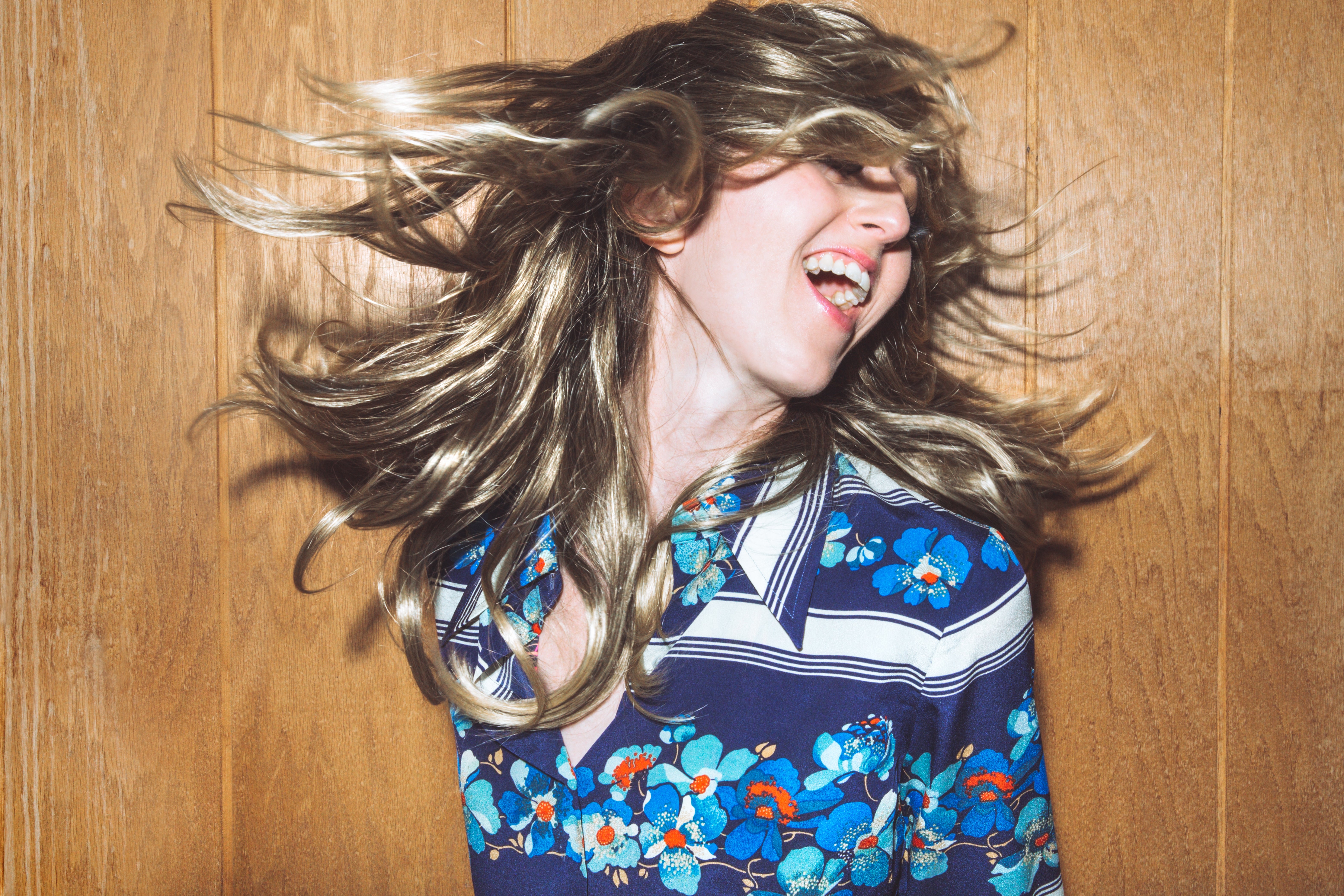 Person joyfully dancing with flowing hair, wearing a floral-patterned shirt against a wooden background
