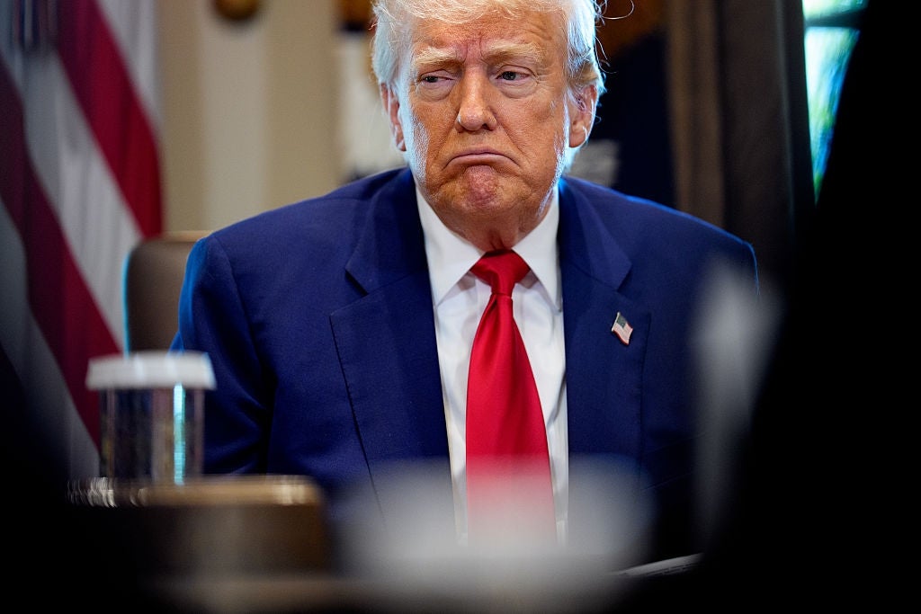 Person in a suit with a red tie sits pensively, appearing thoughtful. An American flag is partially visible in the background