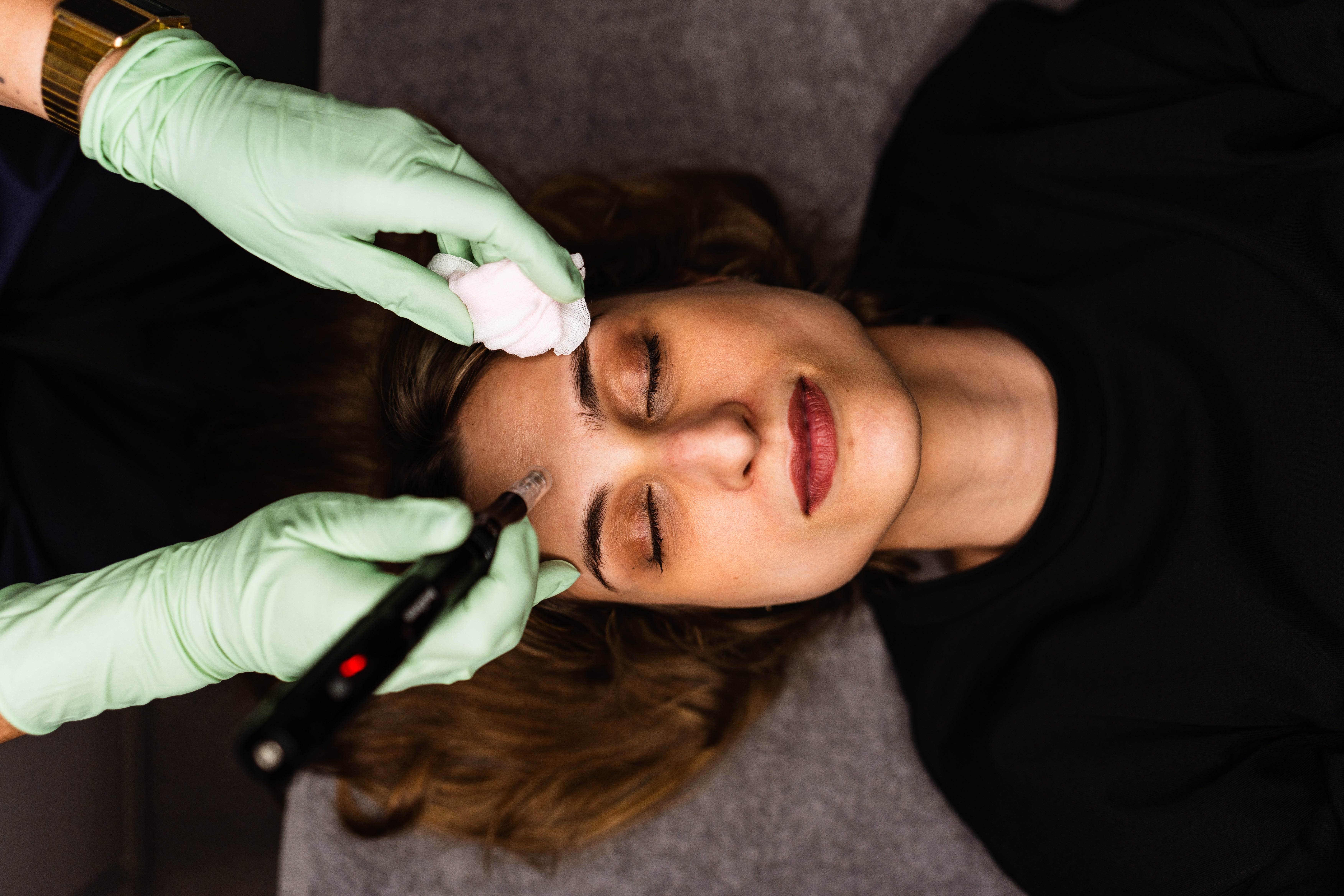 Person lying down receiving a beauty or skincare treatment on their eyebrows, technician wearing gloves holding tools nearby