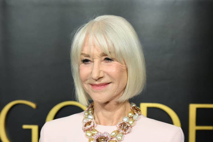 Person with short white hair on red carpet, wearing white top with a statement necklace featuring large pearls and floral accents