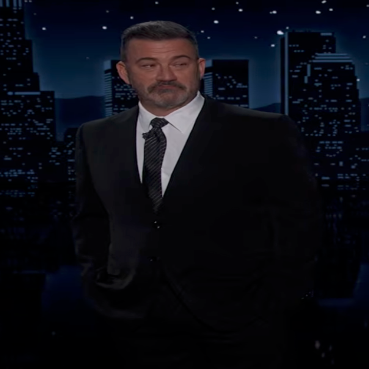 Man in a suit stands against a city skyline backdrop with a night sky, appearing to host a television show