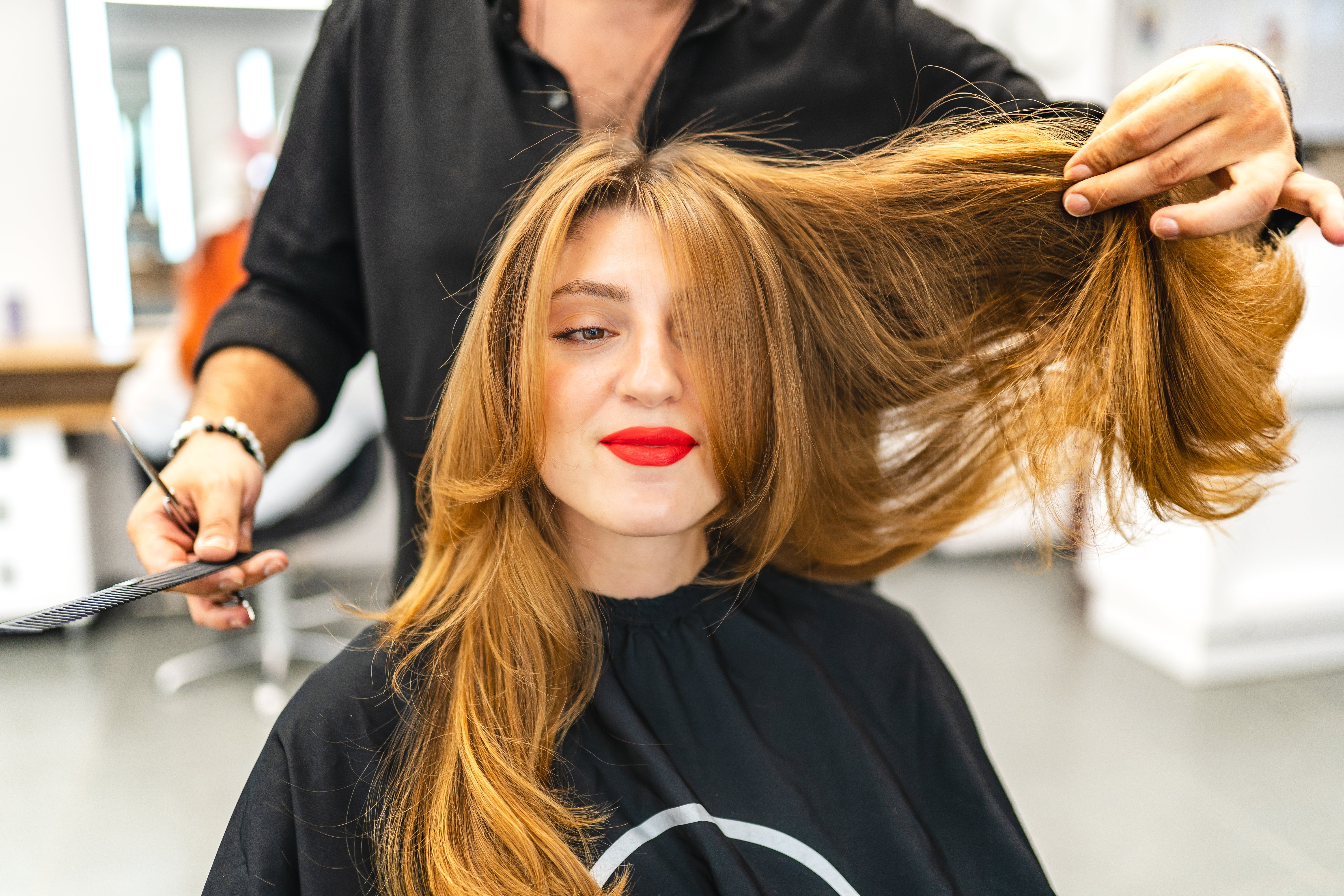 Person with long hair is in a salon. A stylist prepares to cut their hair. The person smiles, wearing a salon cape