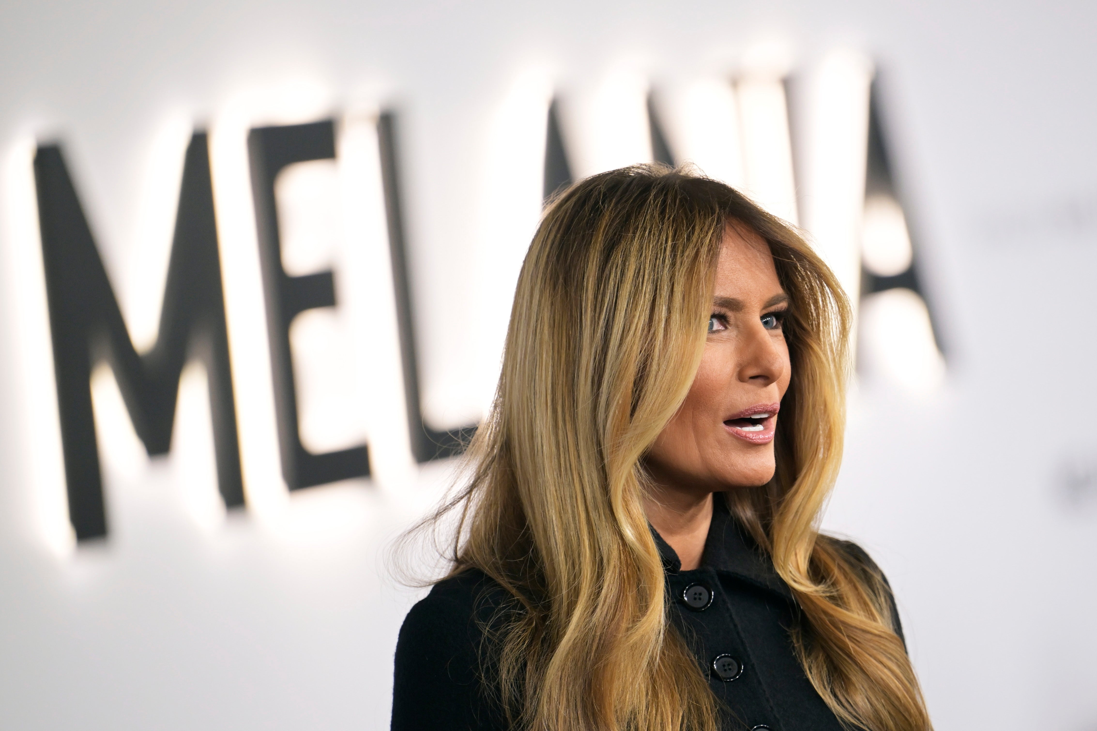 Person with long hair, wearing a stylish coat, speaking in front of a wall with large letters