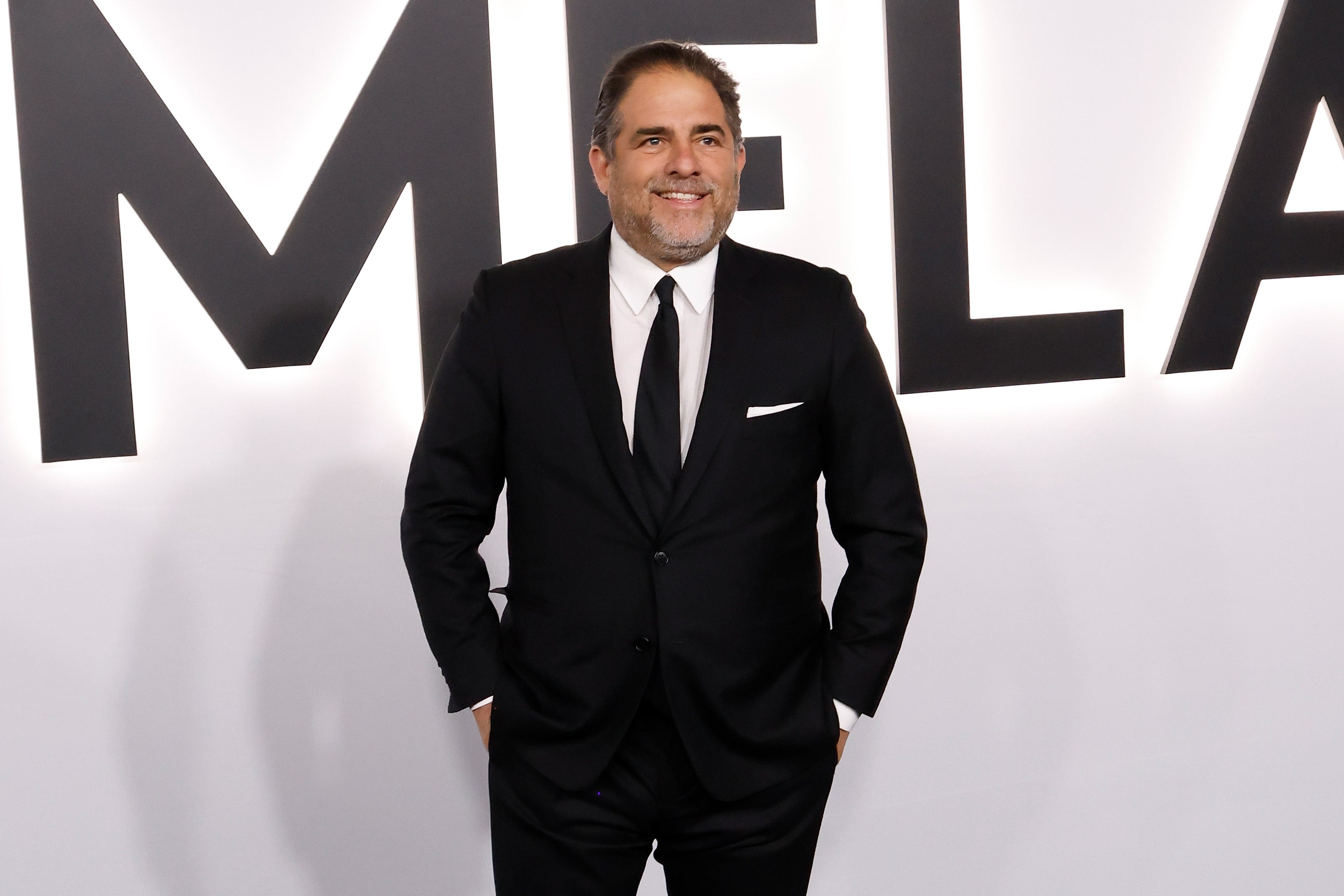 Person in a classic black suit and tie poses confidently at a media event backdrop