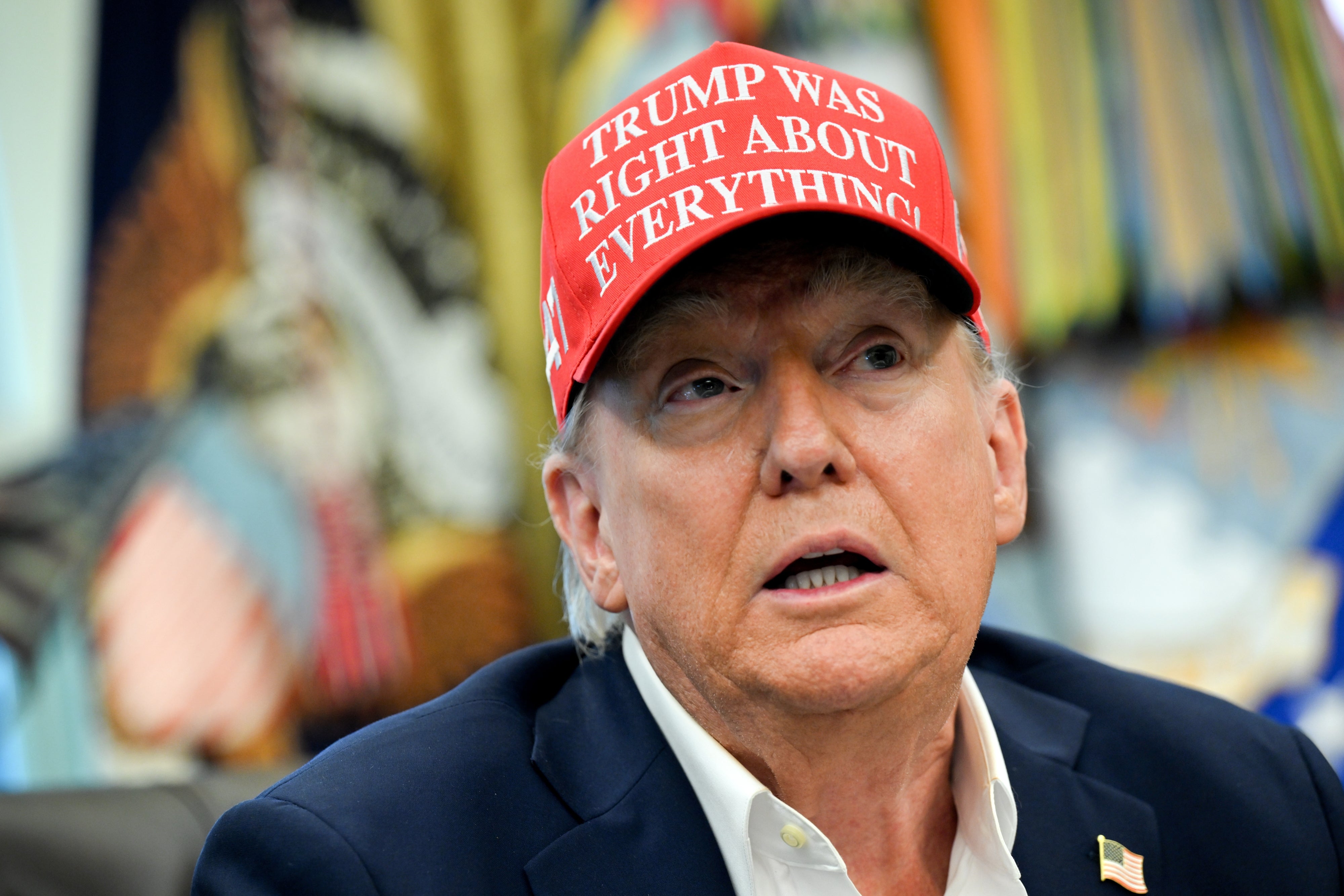 A idiosyncratic   wearing a reddish  headdress  with the substance   "Trump Was Right About Everything!" sits indoors, wearing a suit   overgarment   implicit    a collared shirt