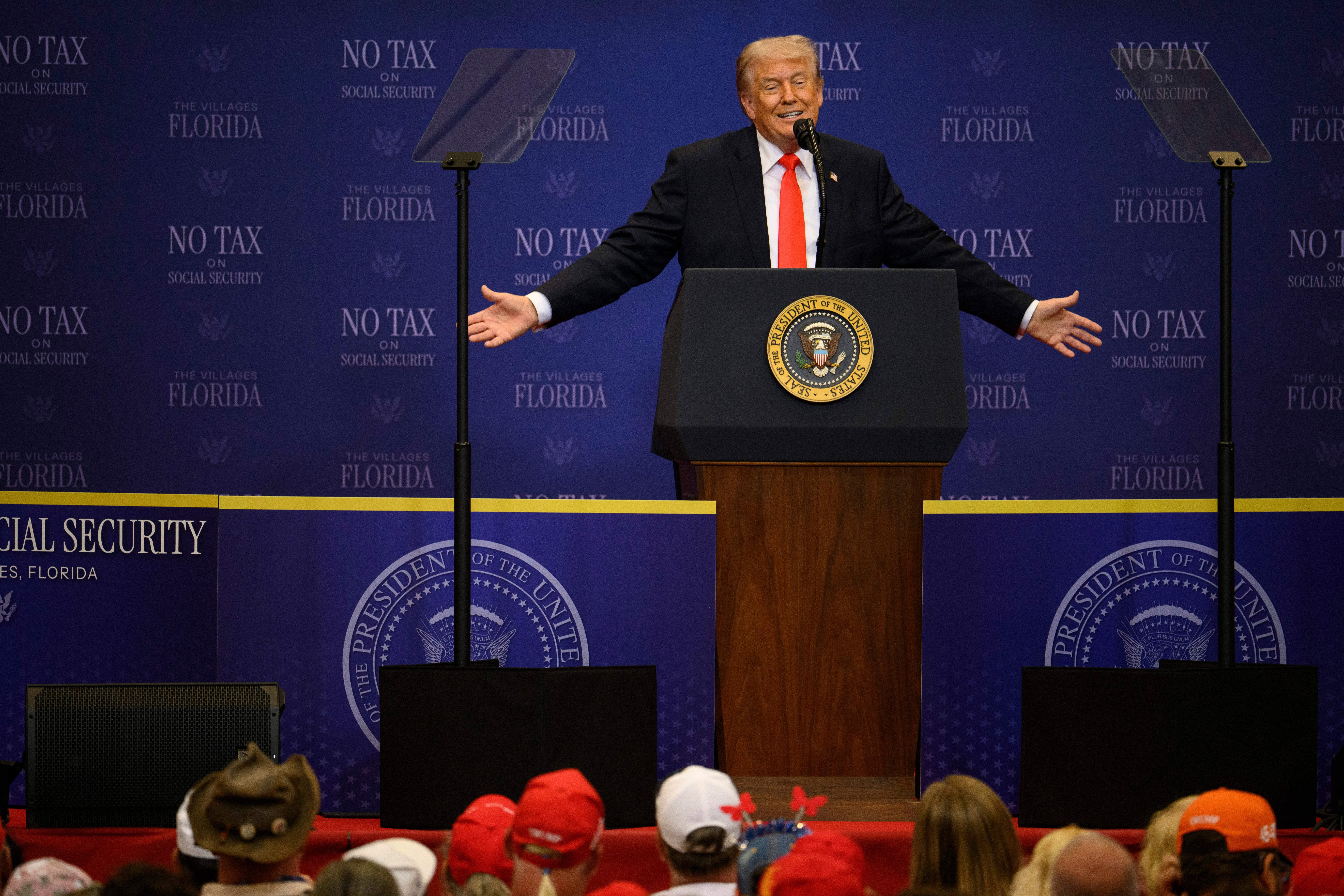 A antheral   speaks astatine  a podium with statesmanlike  seal, arms unfastened  wide, astatine  an lawsuit   with an audience. The backdrop displays "No Tax connected  Social Security."