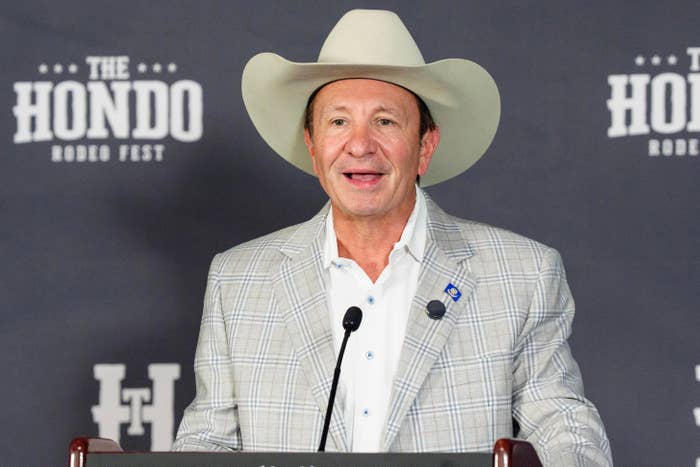 Individual in a cowboy hat and plaid jacket speaks at a podium with "The Hondo Rodeo Fest" banner behind