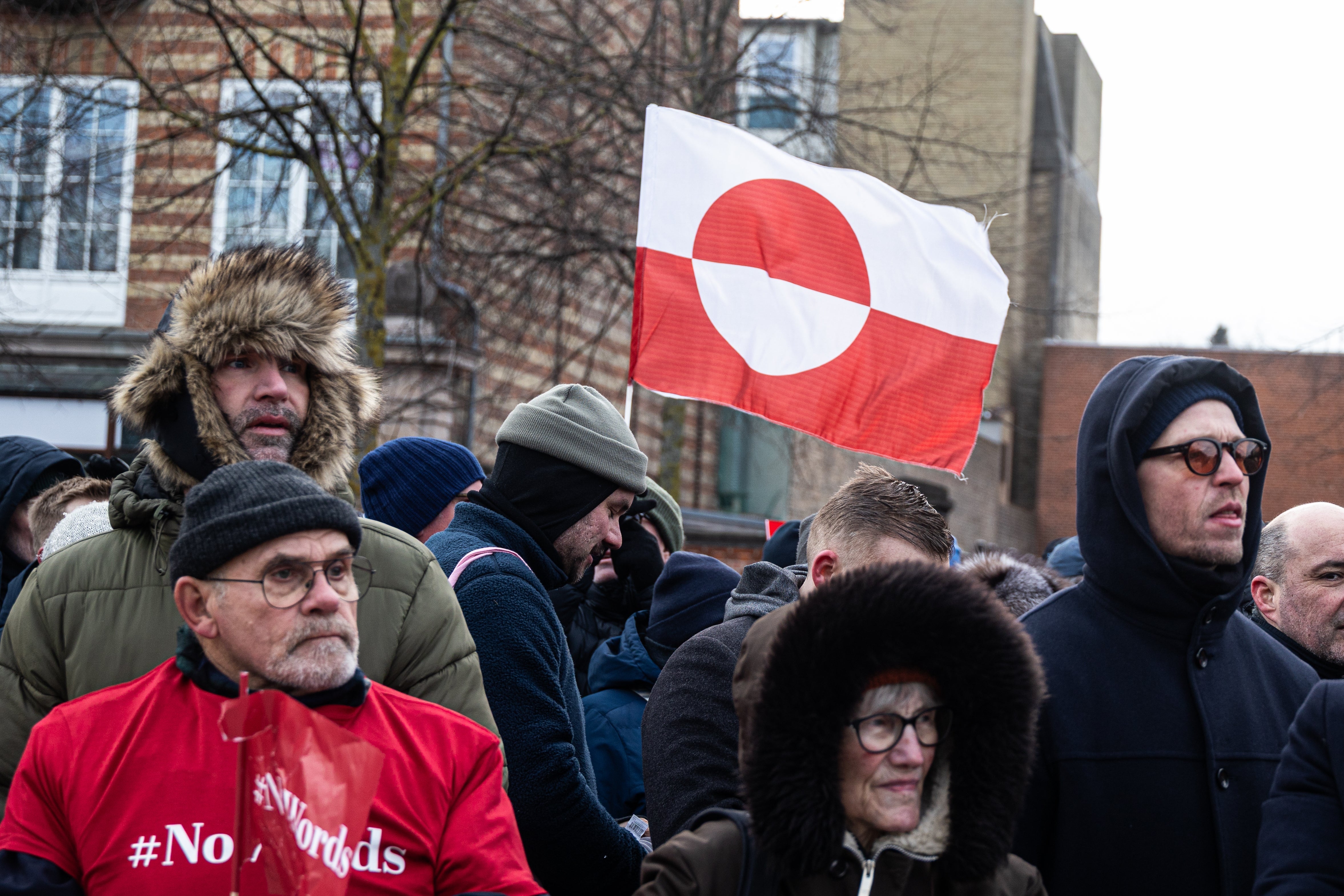 People at an outdoor gathering, bundled in winter clothing, with a flag featuring a red circle and cross in the background