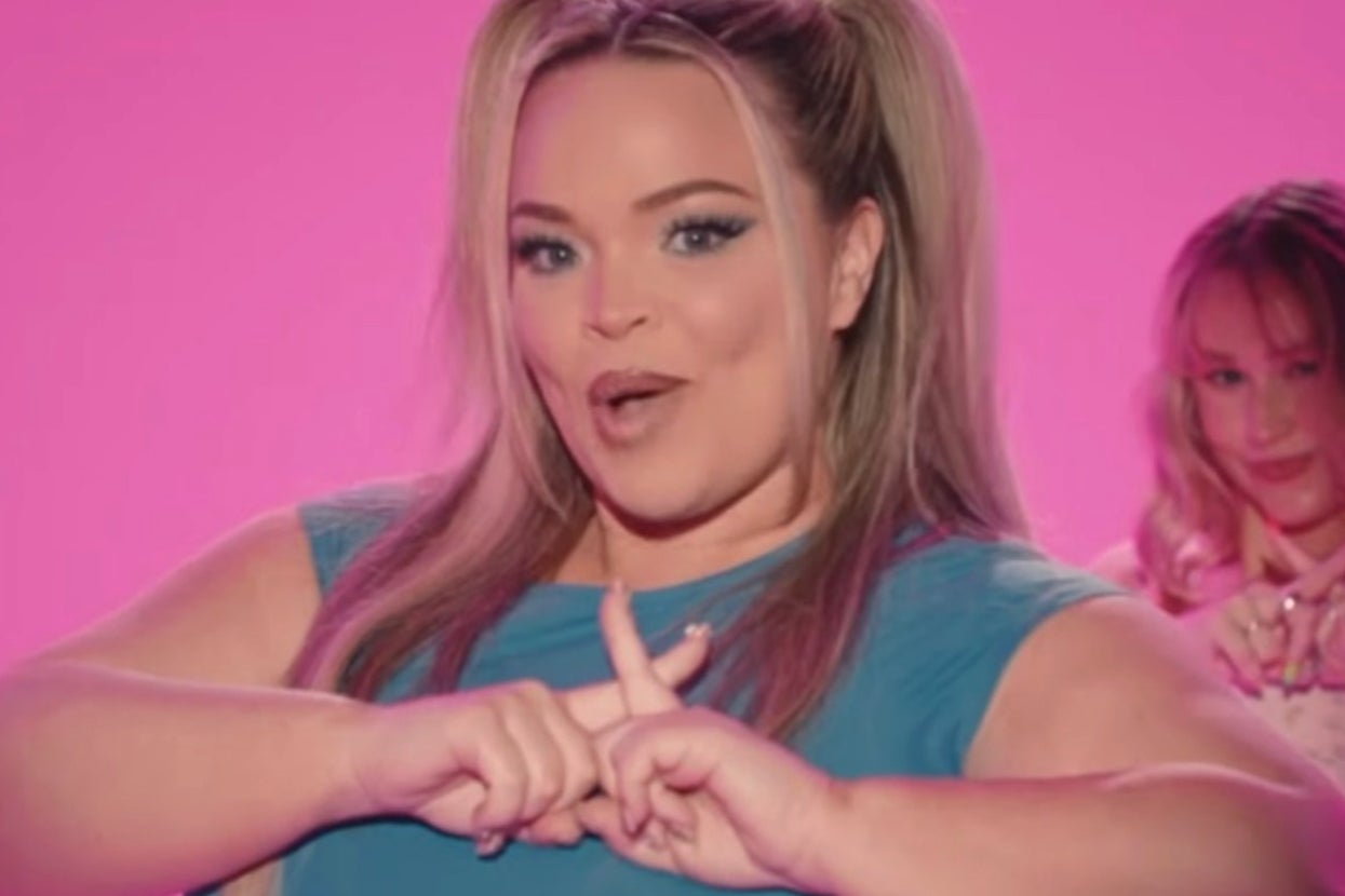 A person with long hair styled in a high ponytail wears a sleeveless top, gesturing in a lively manner in front of a bright background