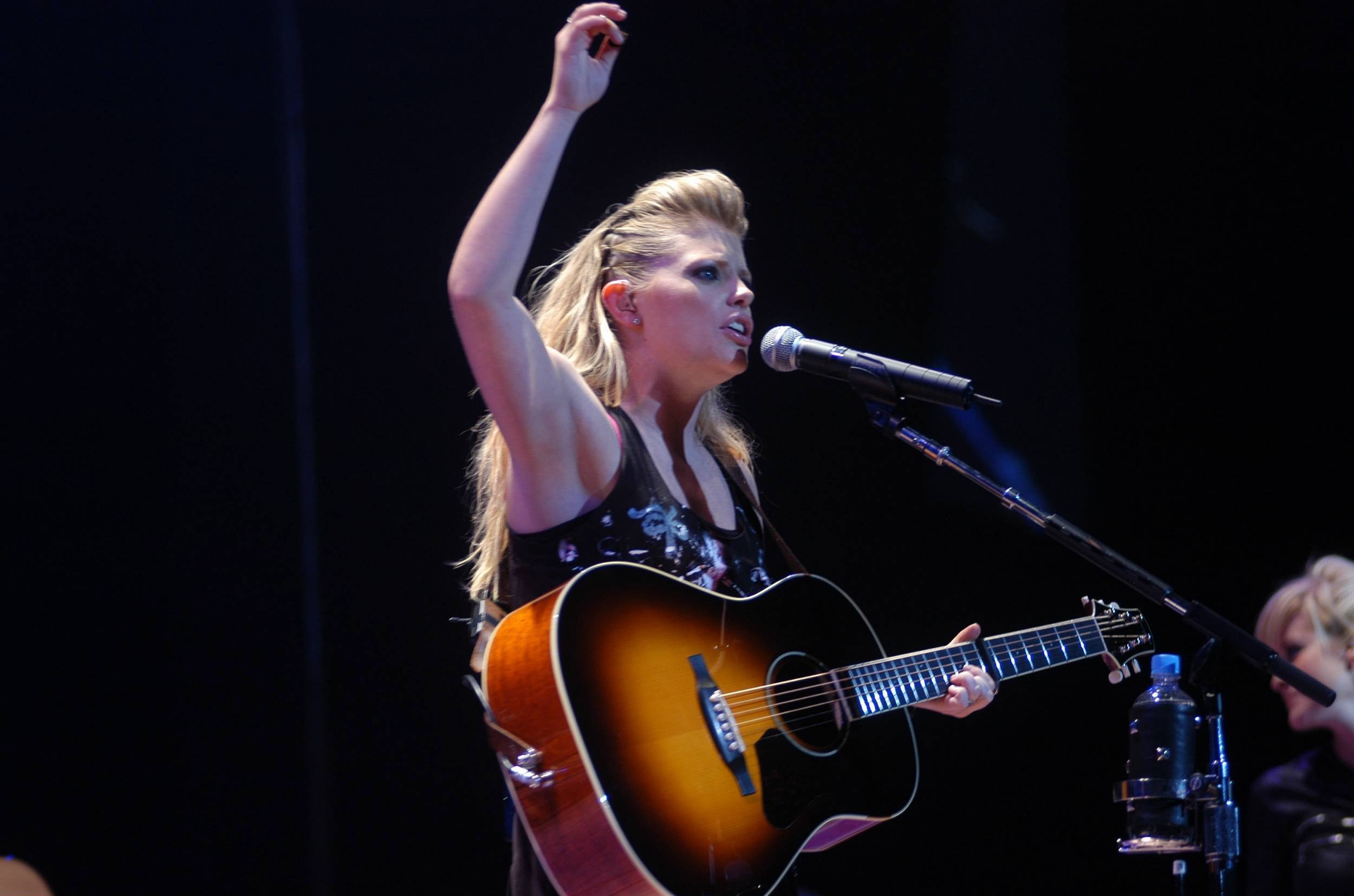 Singer performing on stage with an acoustic guitar, wearing a floral-patterned top, and raising one arm while singing into a microphone
