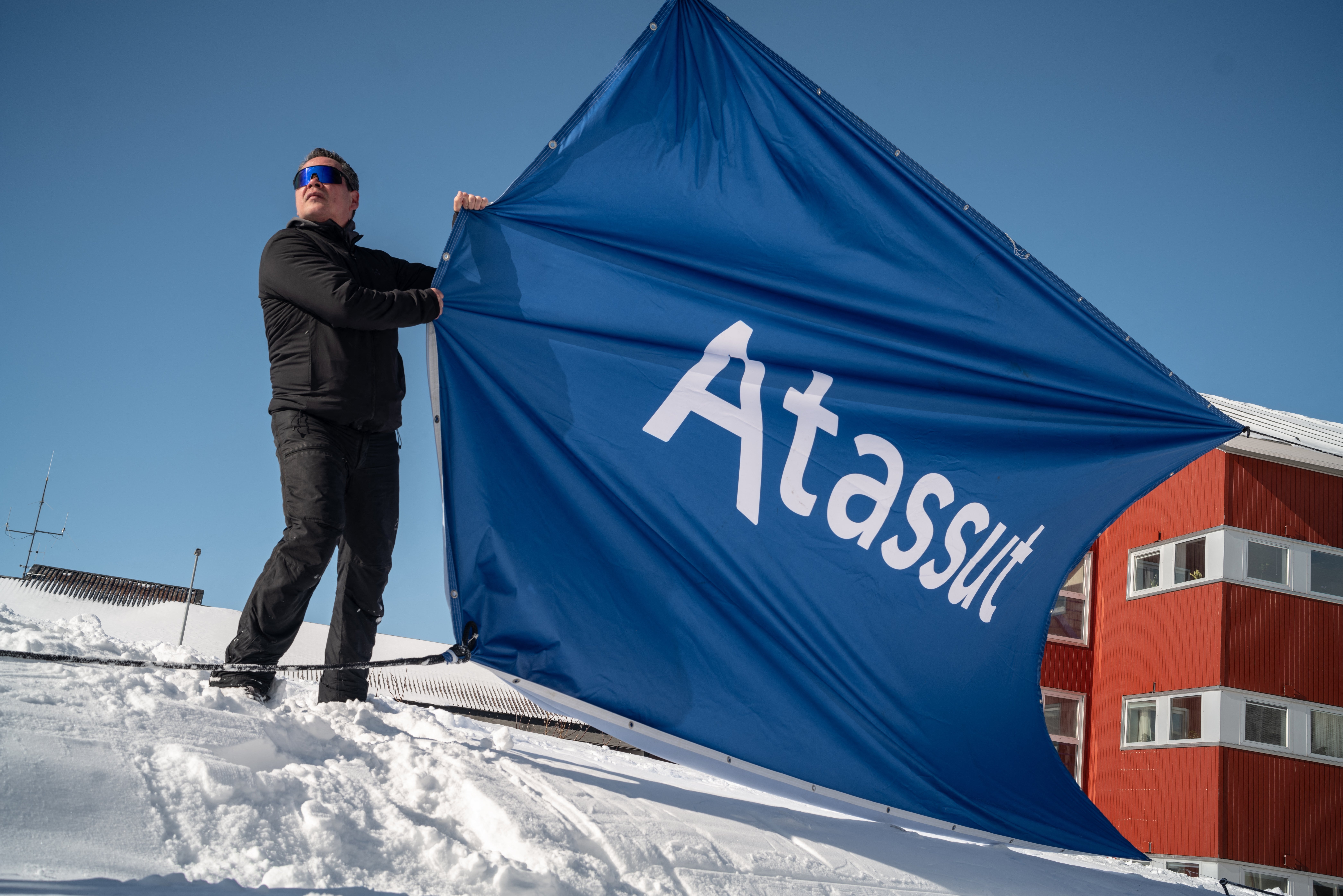 Person holding a large blue banner with "Atassut" text on a snowy rooftop. The background features a red building