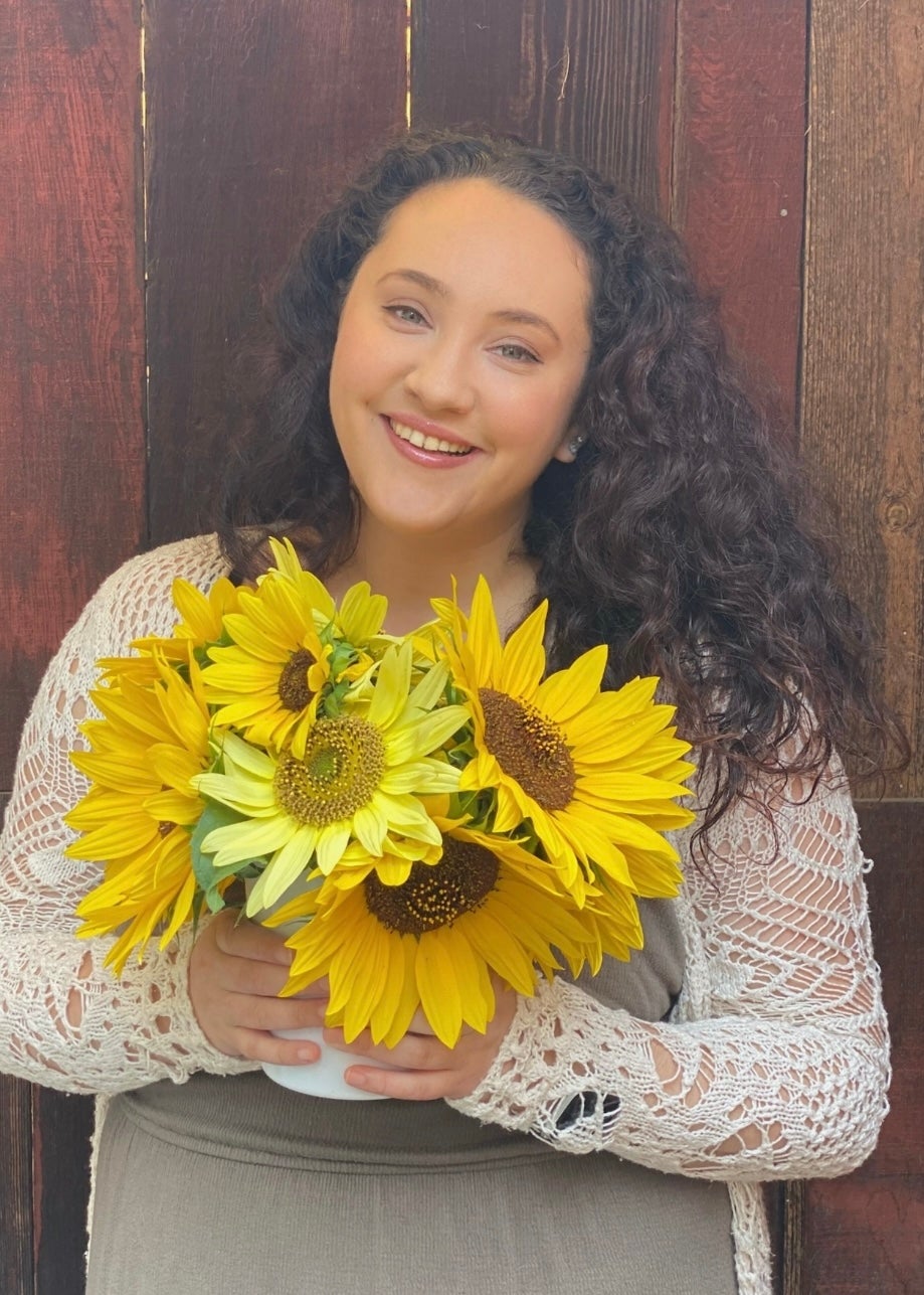 The author is smiling while holding a bouquet of sunflowers, wearing a knit cardigan over a dress