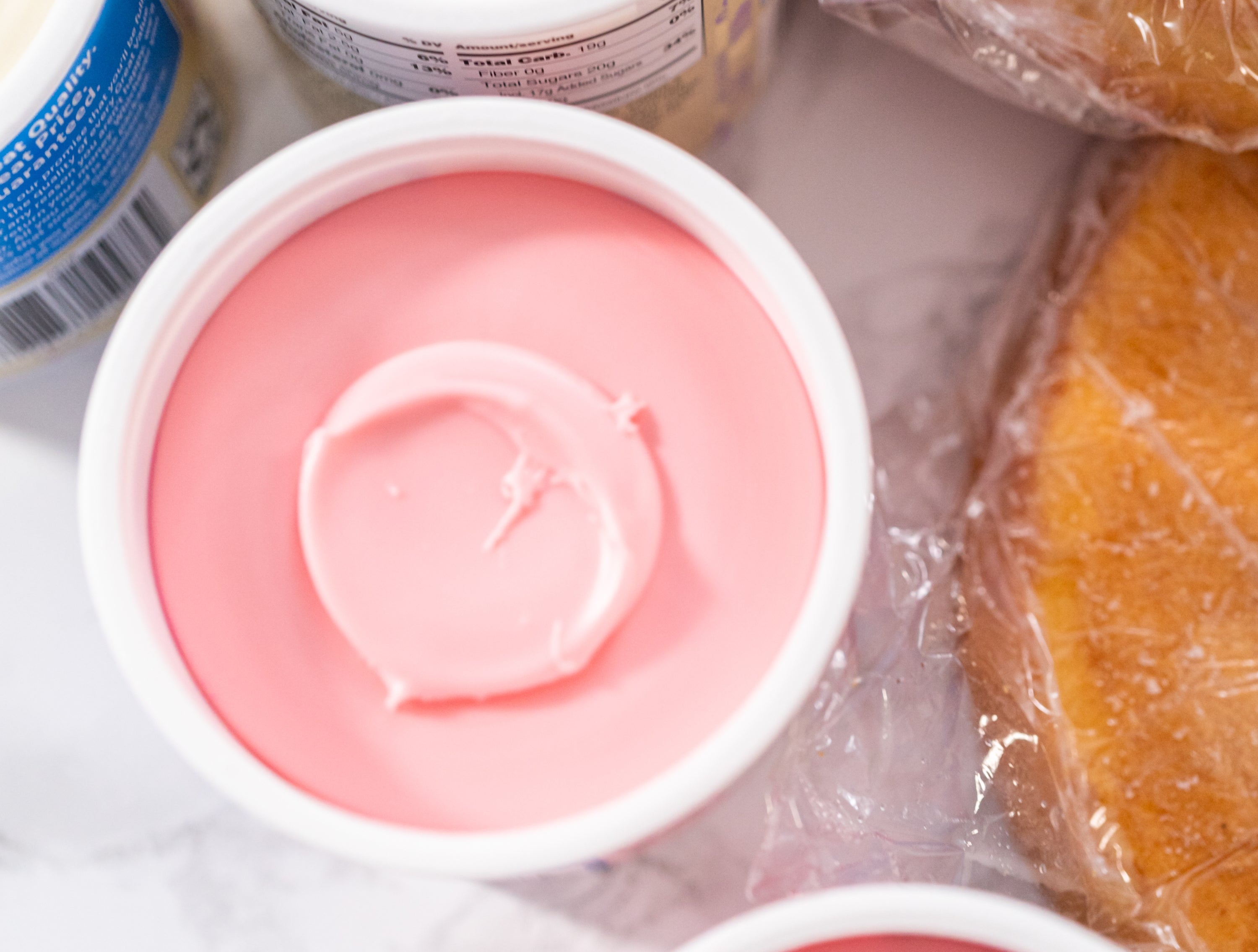 Open containers of pink and white frosting beside packaged buns on a counter