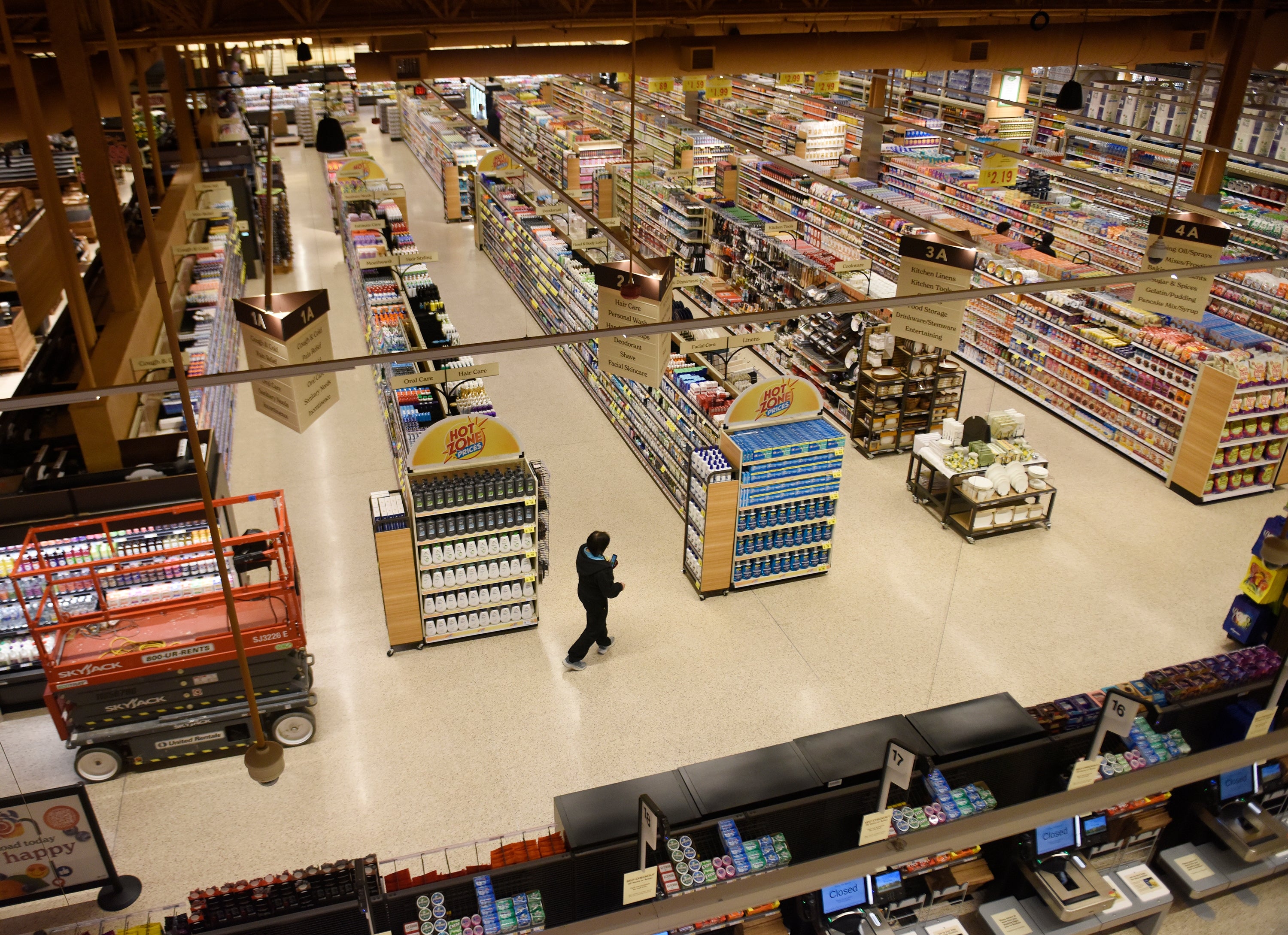 Person shopping in a large grocery store with stocked shelves, viewed from above. Shopping carts and products fill the aisles