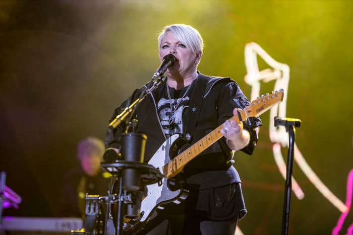 Person performing on stage with an electric guitar, dressed in a black outfit. Microphone and music equipment visible