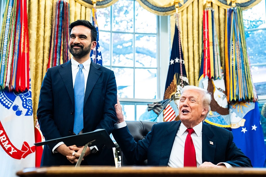 Two men in formal attire are in an official-looking room. One man is seated with a pen, smiling, as the other stands beside him, also smiling