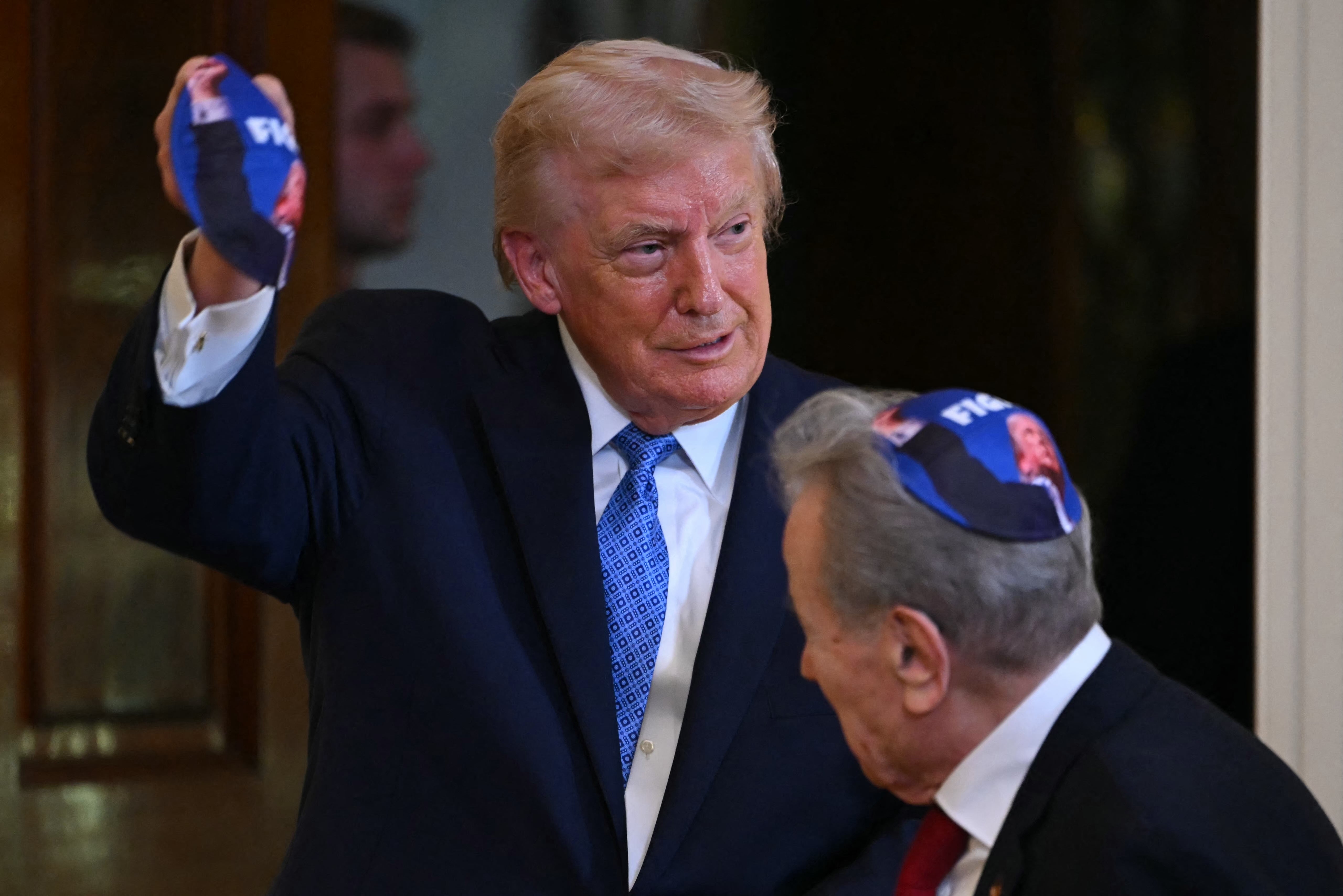 Two people in suits, one holds and wears a yarmulke featuring a printed image, suggesting a public or formal event