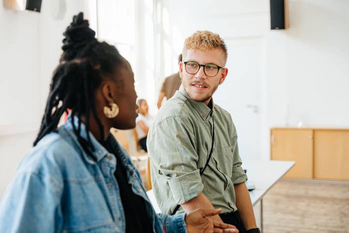 Two people sitting and talking in a room with large windows. One has braided hair and the other has short curly hair and glasses