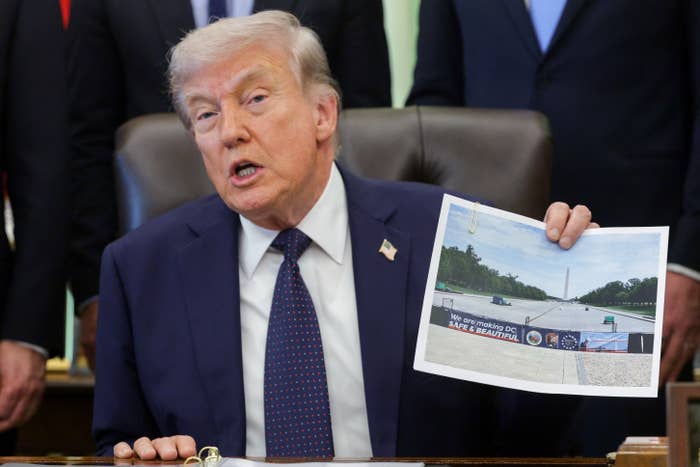 A person in a suit sits at a desk holding a printed image of a monument area while speaking, with people standing behind