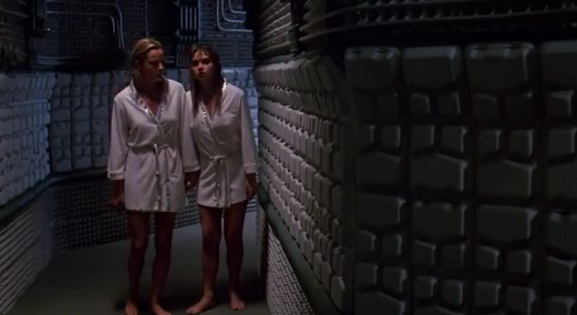 Two people in white robes walk barefoot in a futuristic hallway with metallic walls