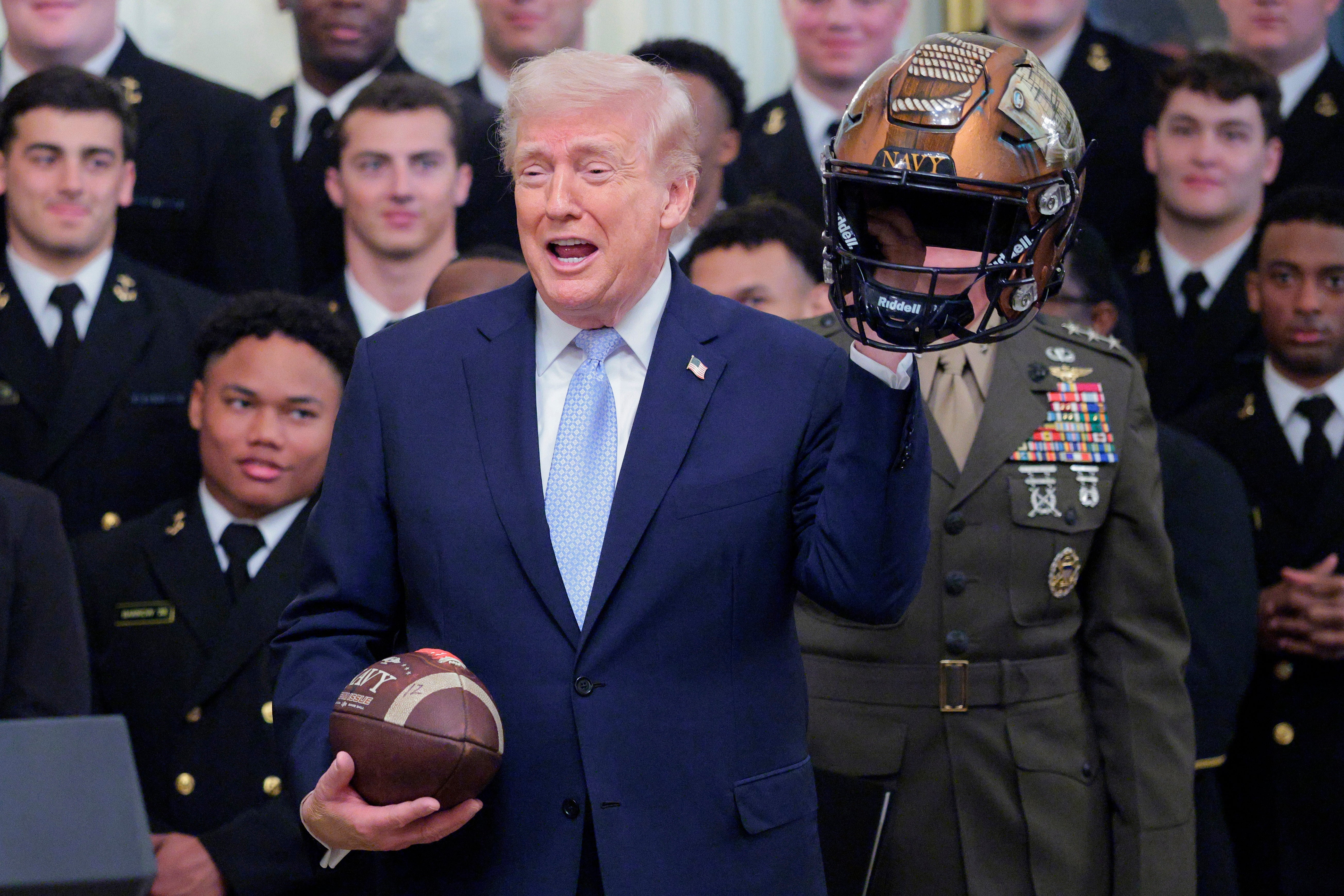 A person in a suit holds a football and gestures while another in uniform holds a helmet. Group of people in background