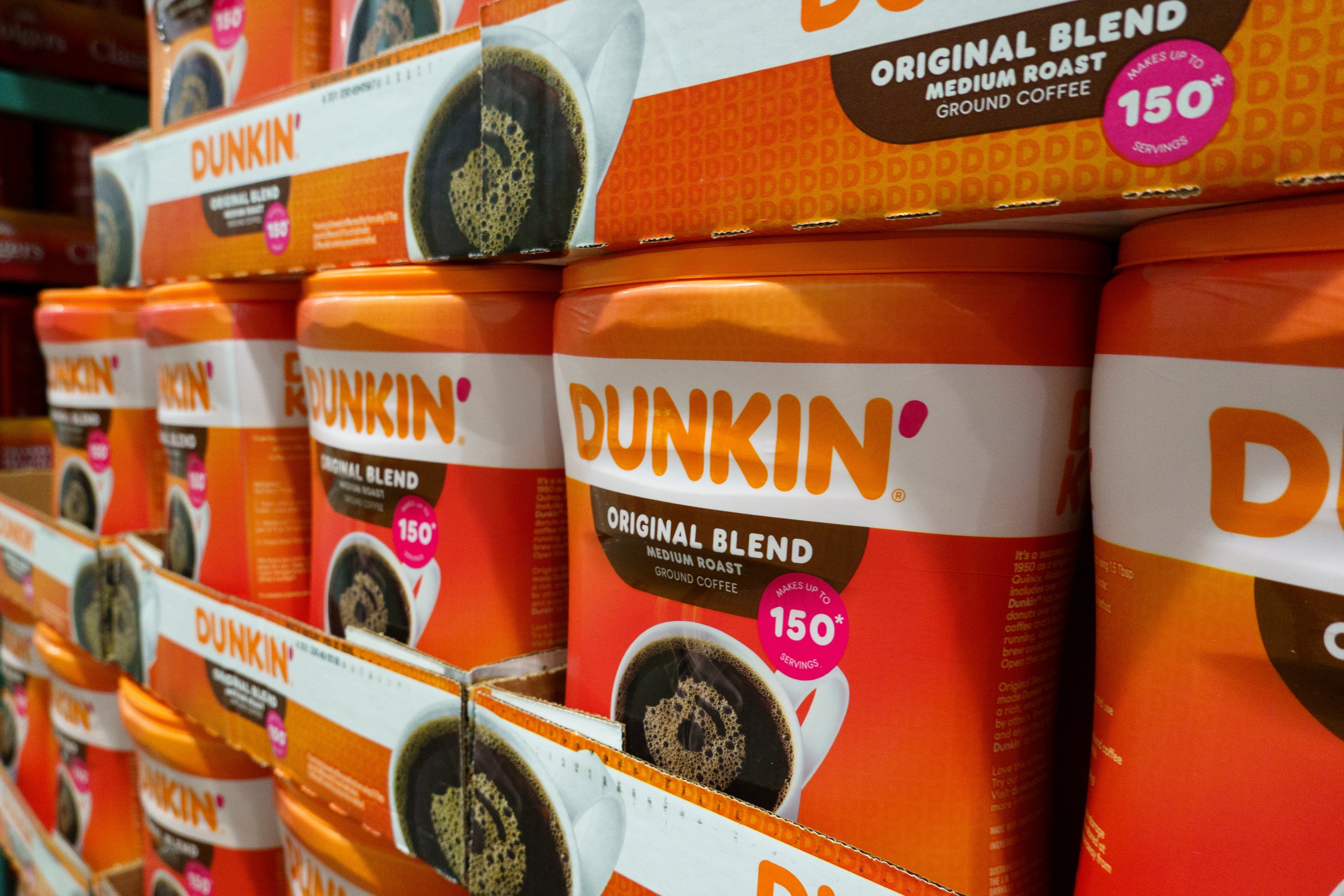 Stacks of Dunkin' Original Blend coffee cans on store shelves, highlighting a "150 cups" label for bulk purchase appeal