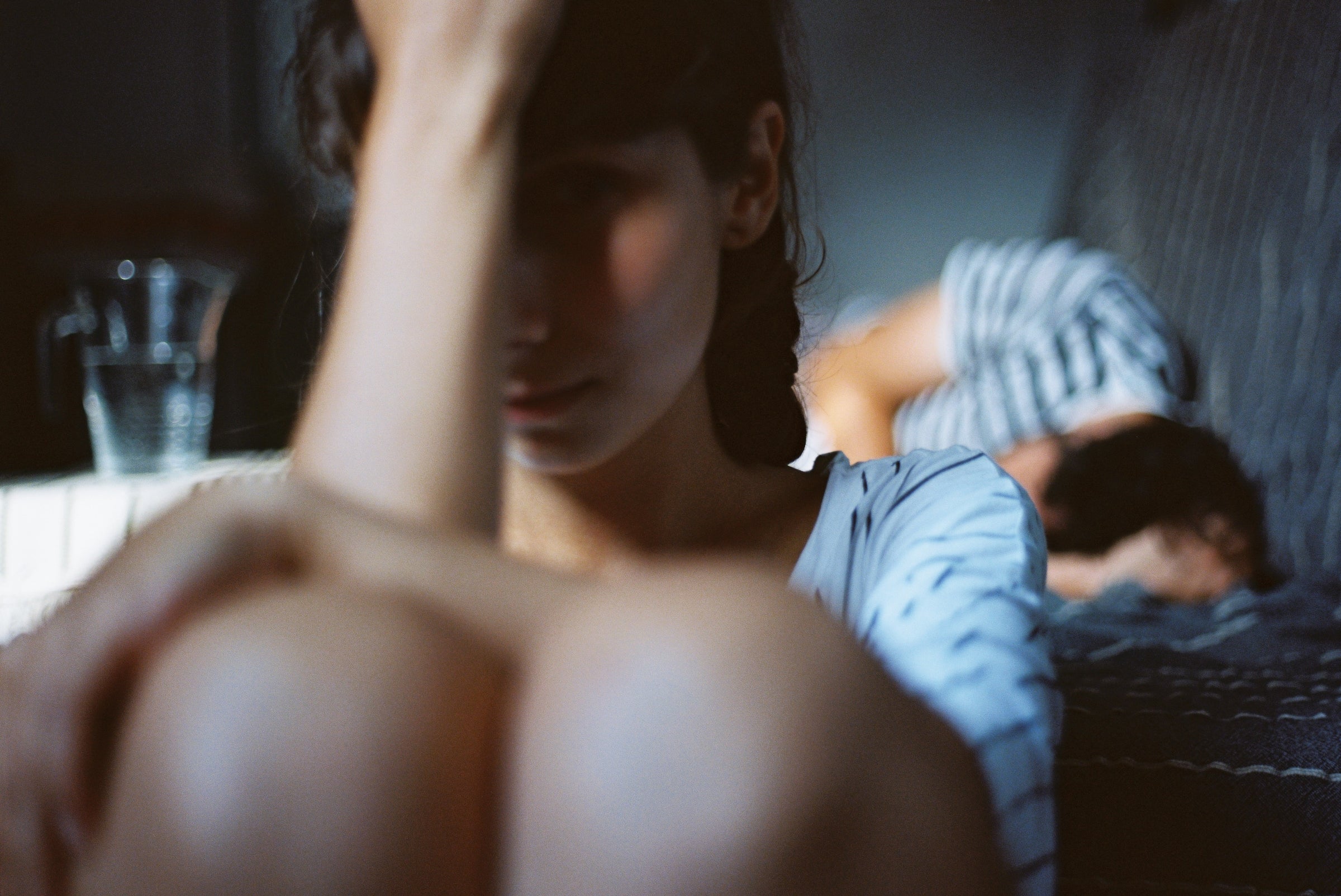 Two people in a dimly lit room, one sitting with a hand on their forehead, the other lying in the background