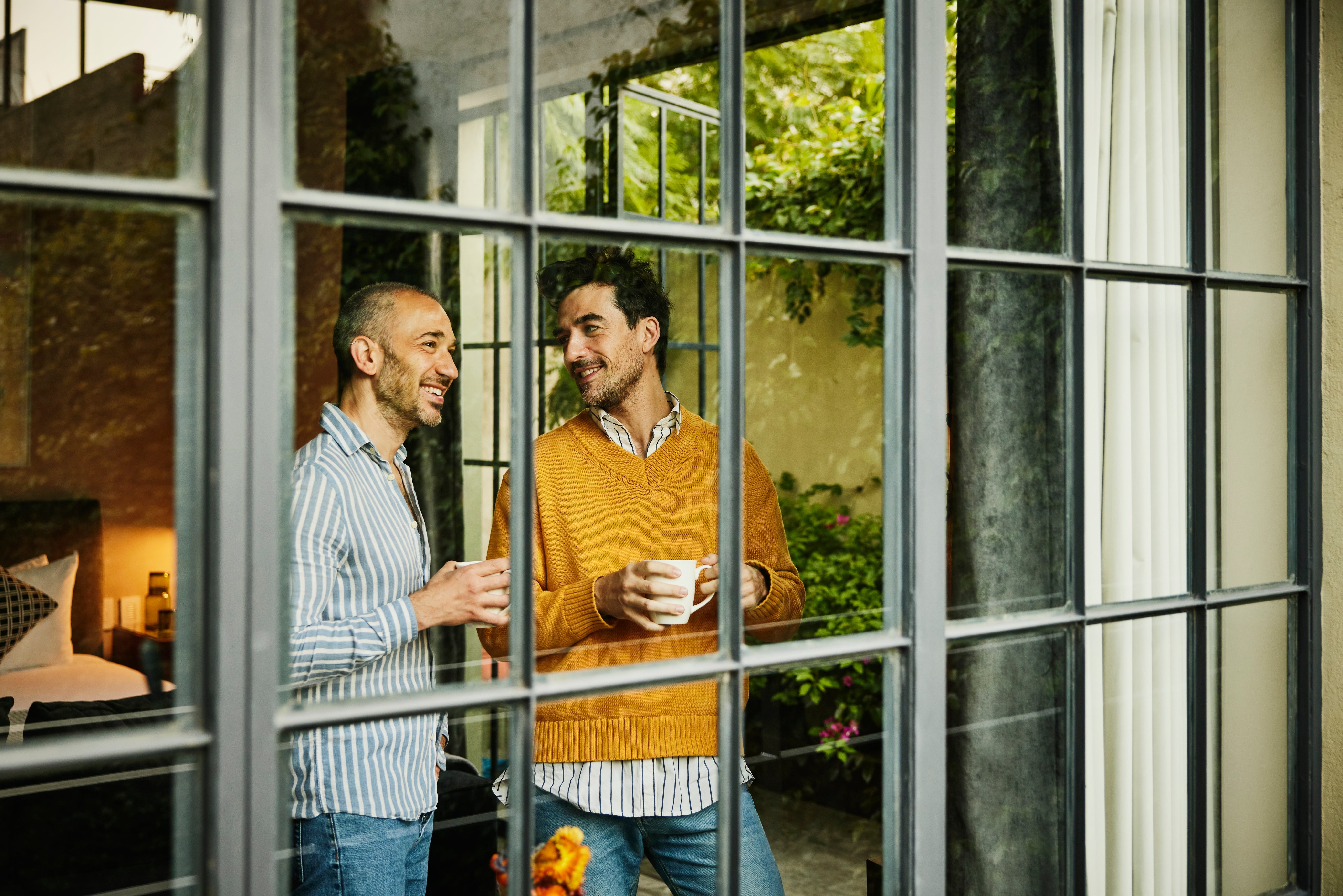 Two men smiling at each other through a window, each holding a coffee cup, in a cozy, relaxed setting