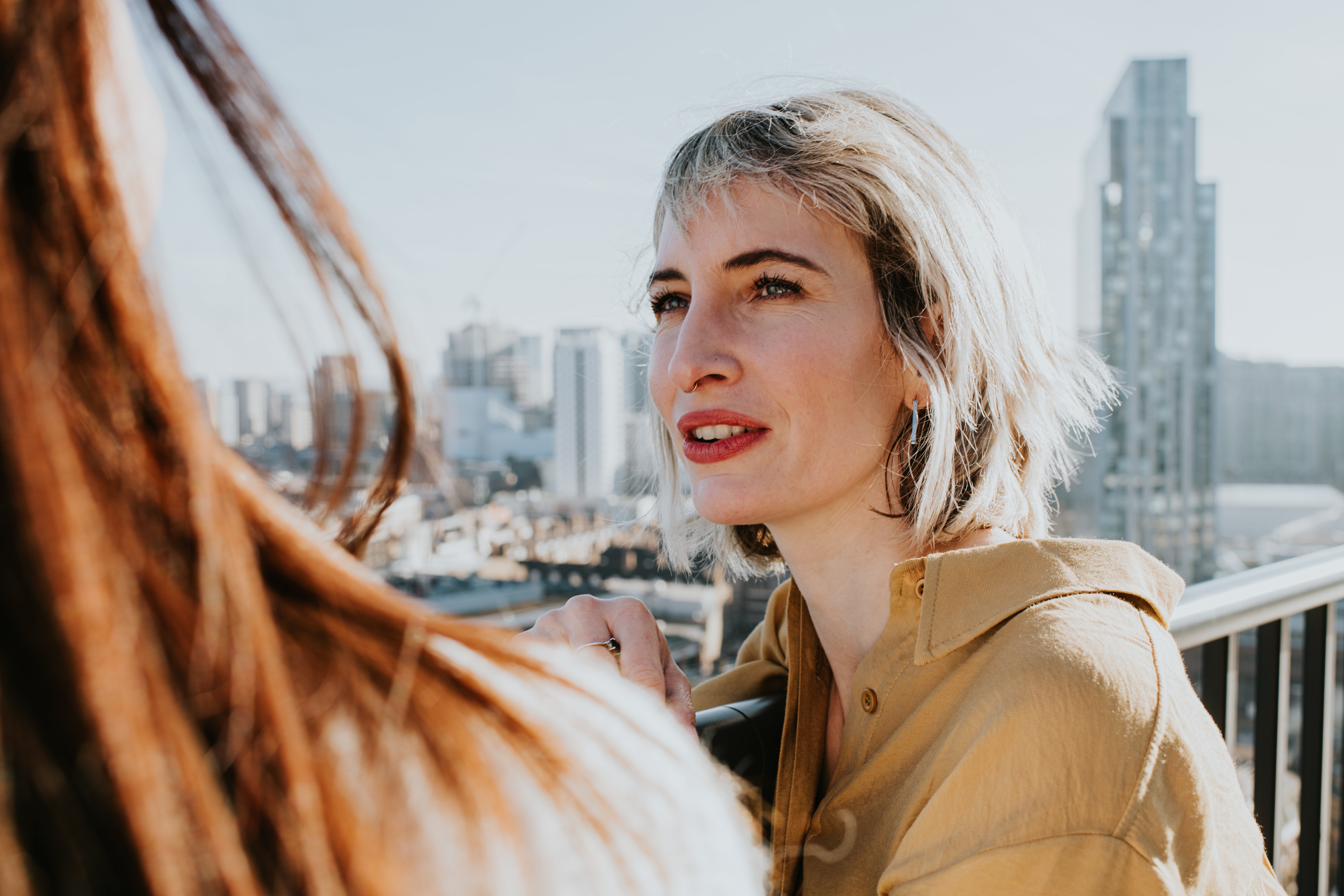 Two people converse on a balcony overlooking a cityscape; one with short hair and gold-toned shirt listens attentively