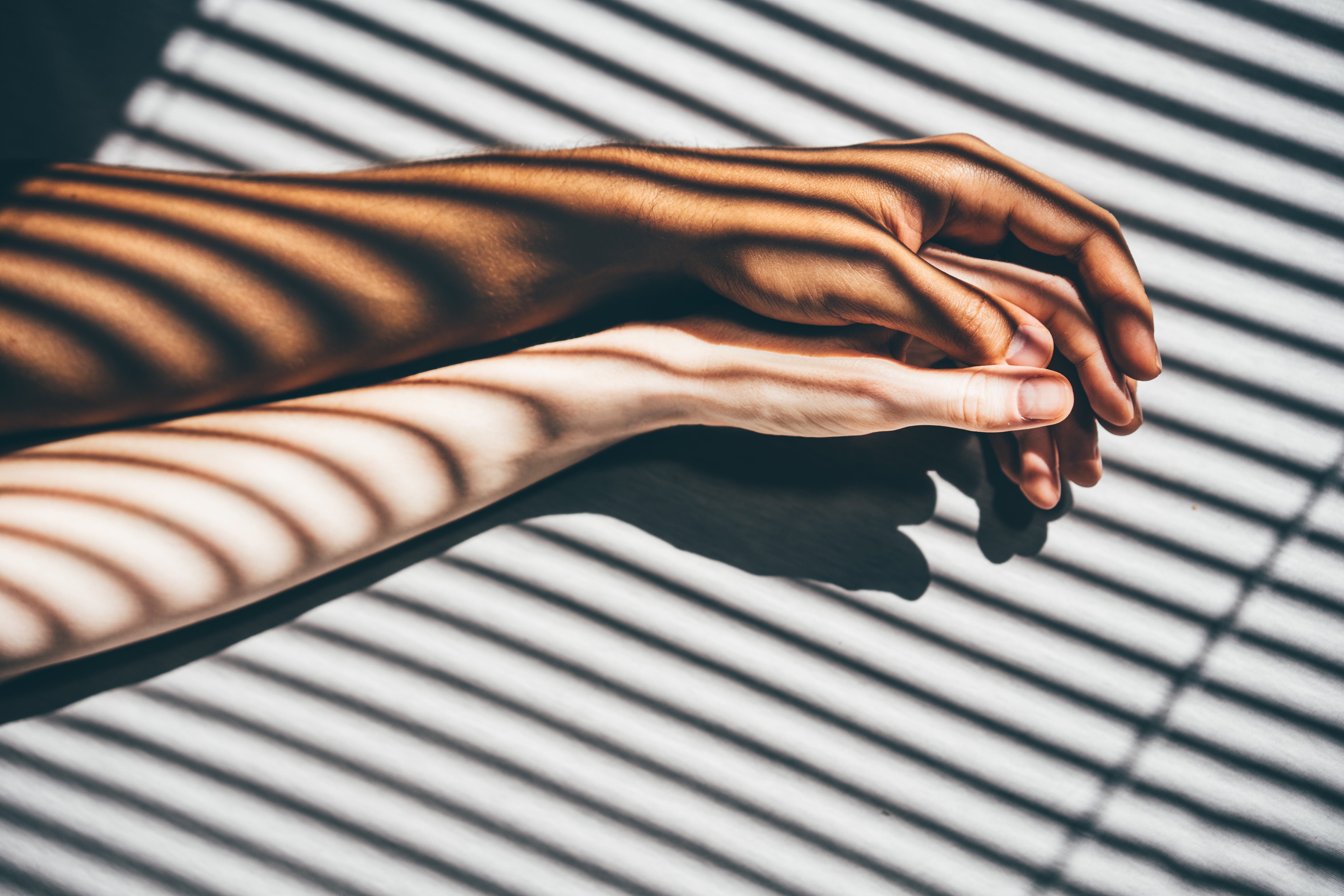 Two hands, one darker and one lighter, gently touch on a surface with striped shadow patterns, suggesting connection and intimacy