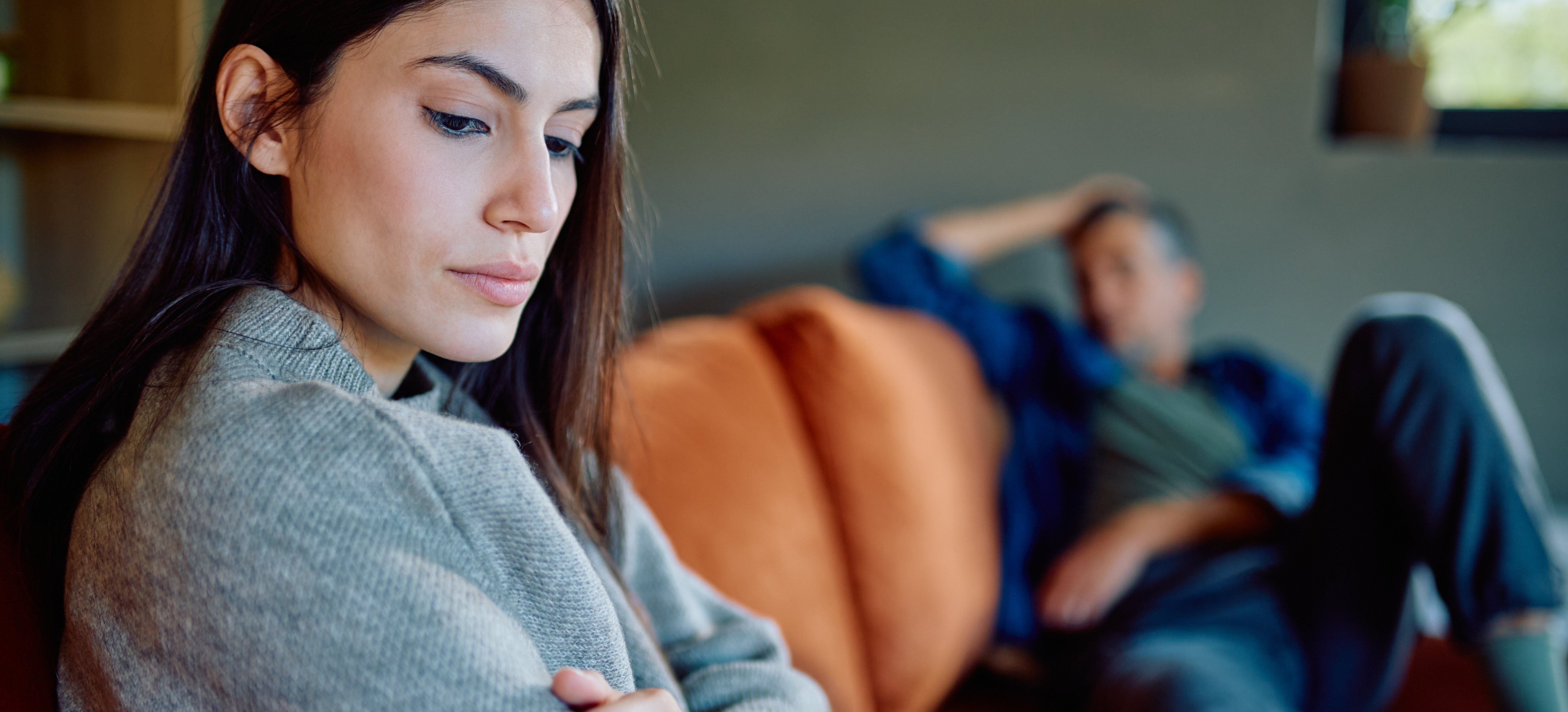 A woman sits on a couch, looking down with a thoughtful expression, while a man in the background appears relaxed but distant