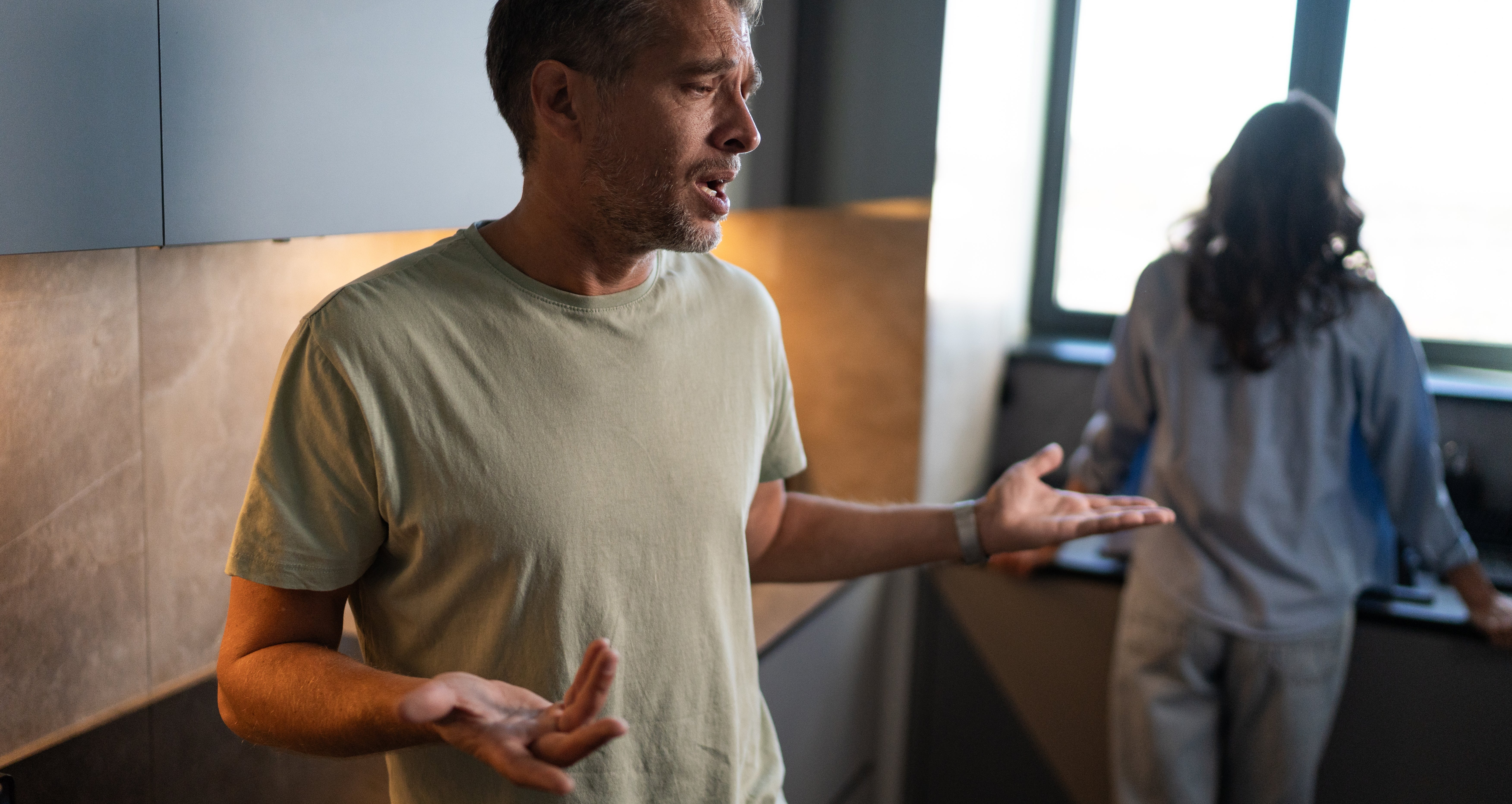 Man in casual attire looks frustrated, gesturing with hands in a kitchen. Woman in background, facing away near window, adds tension to the scene