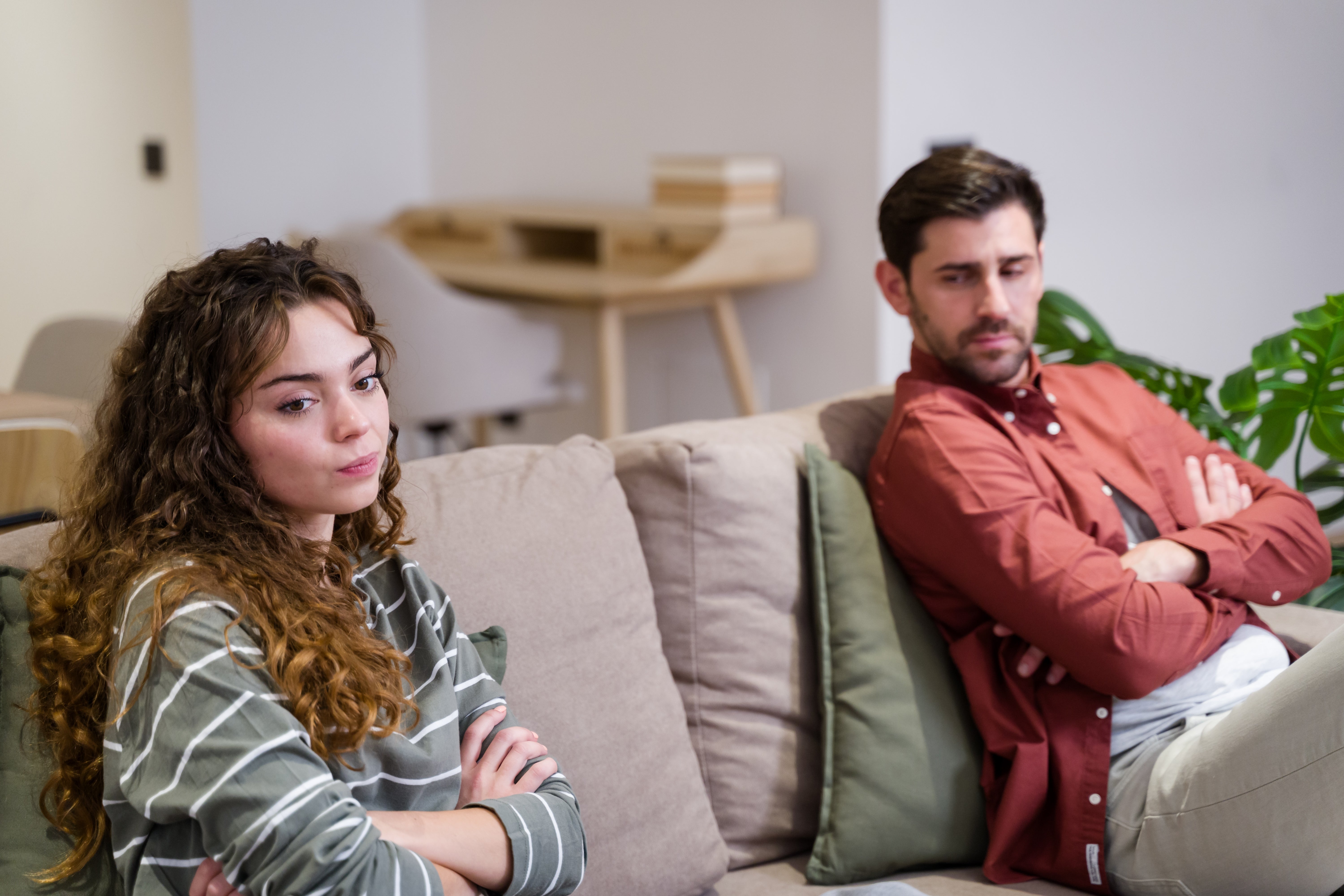 Two people sitting on a couch with arms crossed, looking away from each other, appearing thoughtful or in disagreement