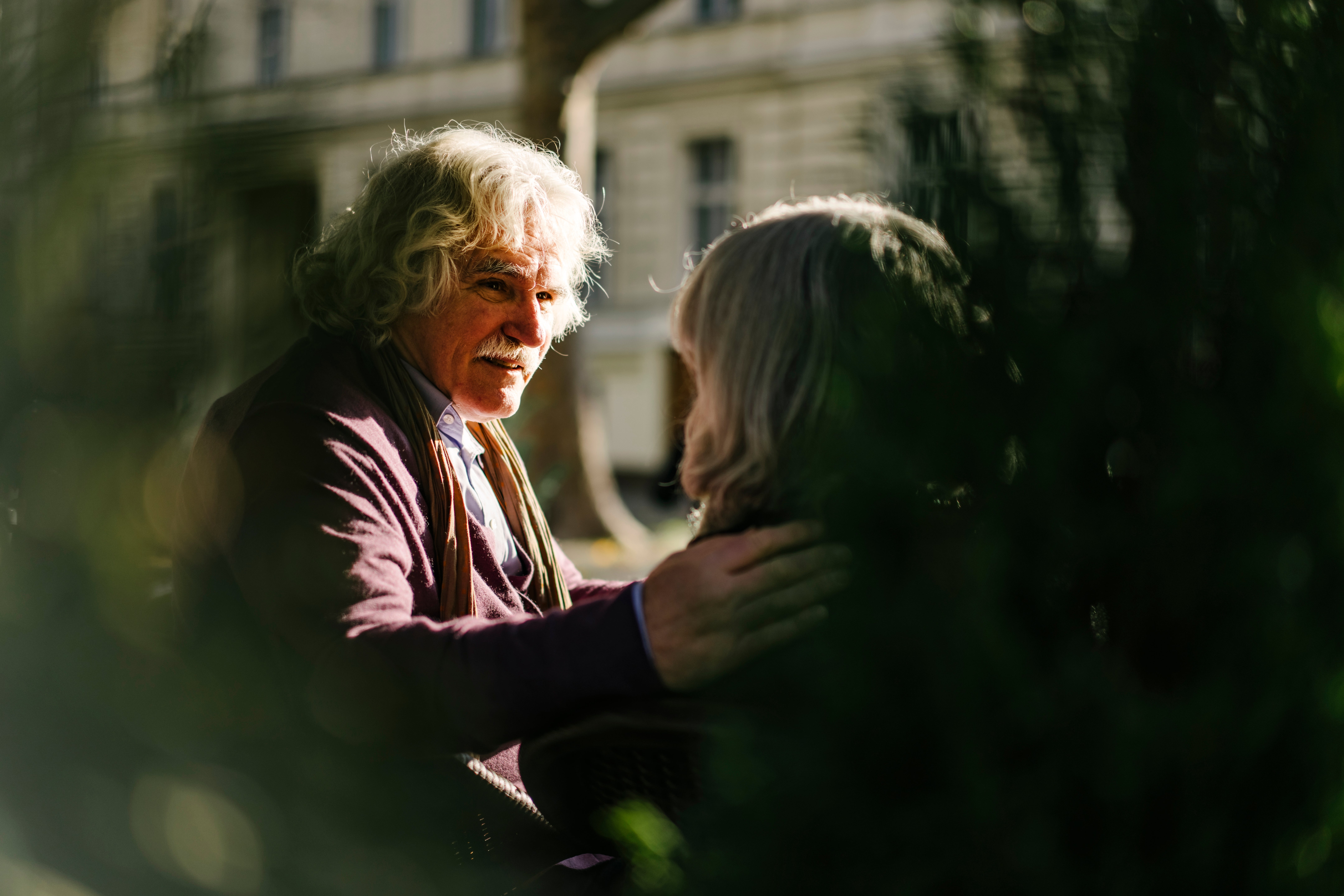 An older couple shares an intimate moment outdoors, with the man gently holding the woman's shoulder while talking