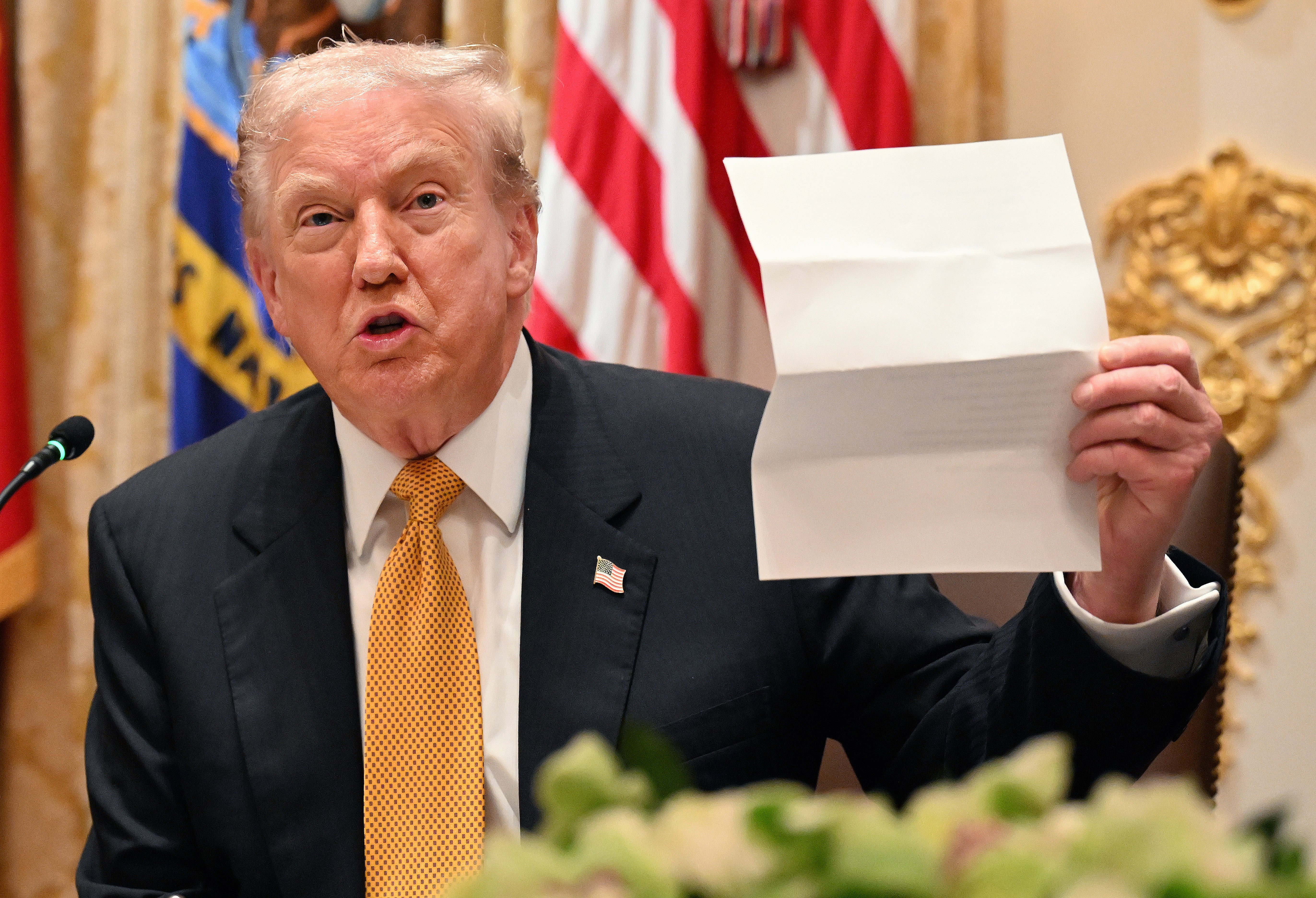A person in a suit holding a sheet of paper at a formal event, with flags in the background