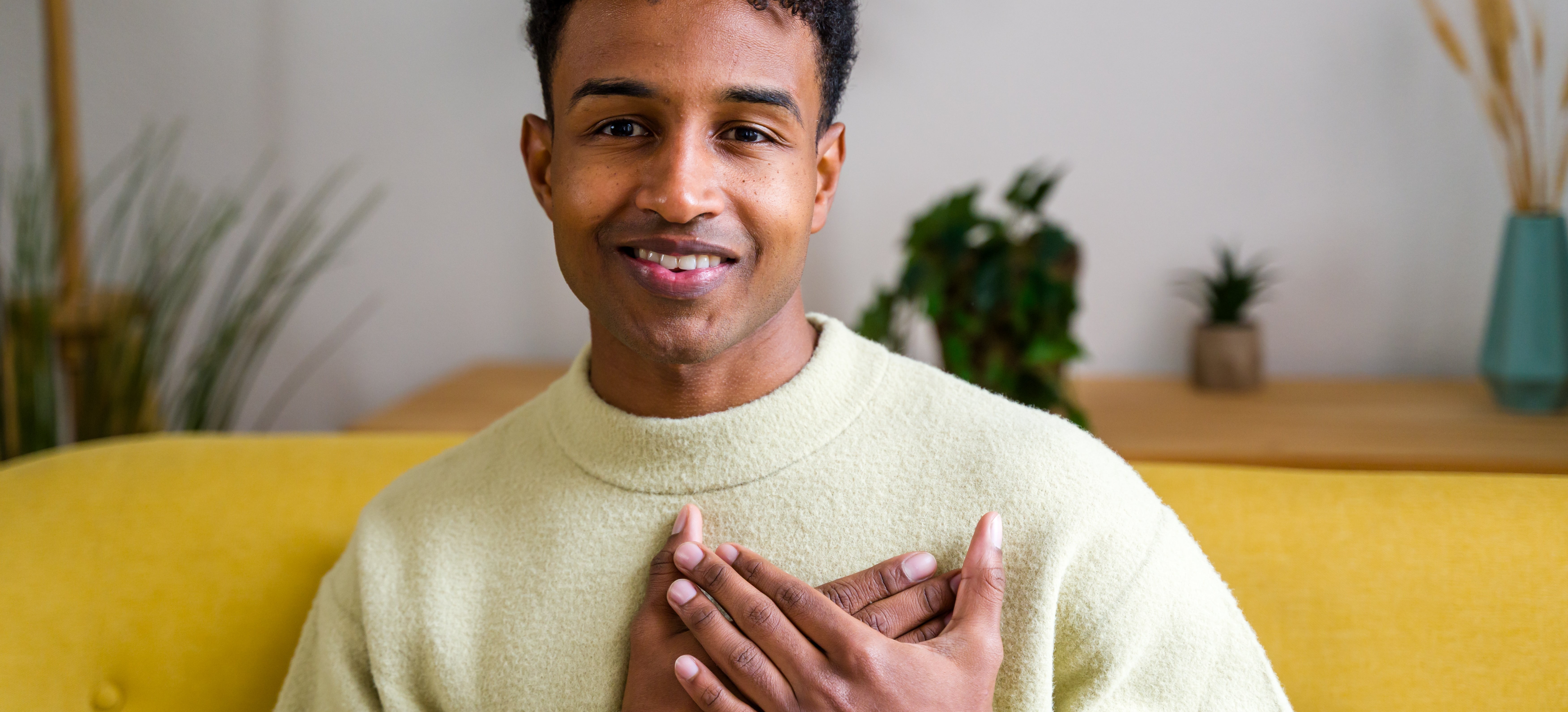 Person smiling warmly with hands over heart, wearing a cozy sweater, sitting on a couch. They convey a sense of affection and sincerity