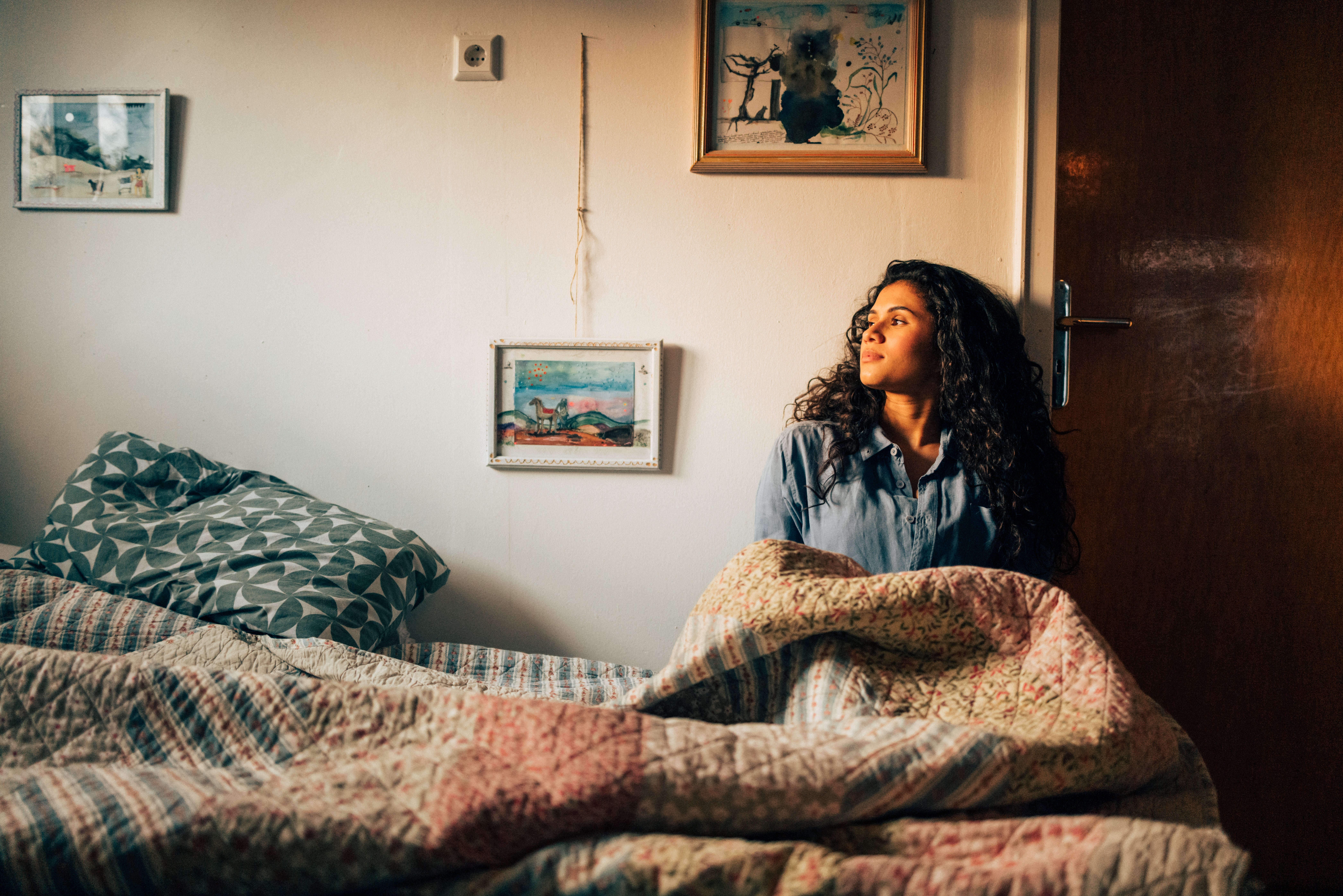 A person with curly hair sits on a bed, looking thoughtfully out a window in a cozy, art-decorated bedroom