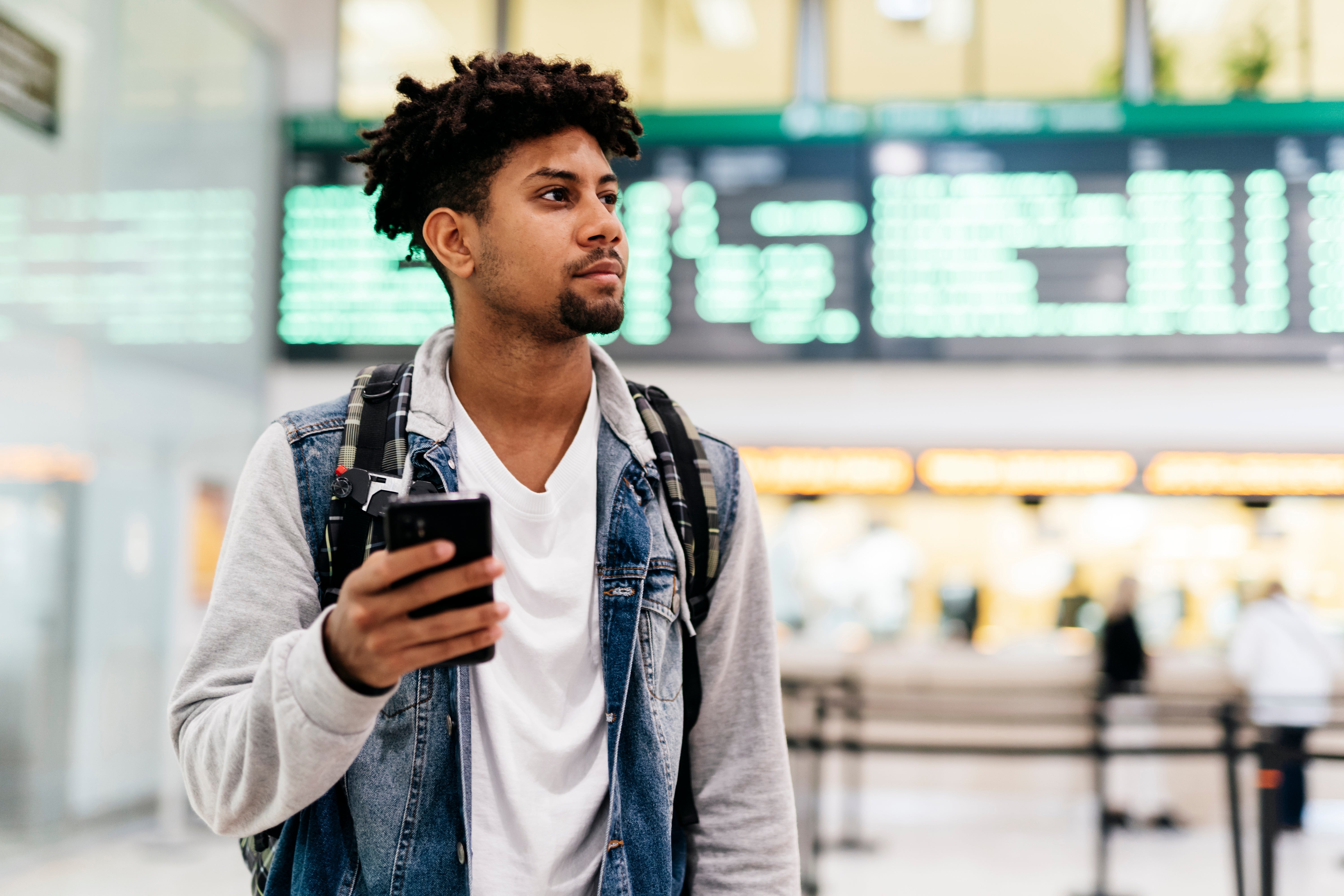 Young man with a backpack holding a phone, standing in an airport terminal with digital flight information displays in the background