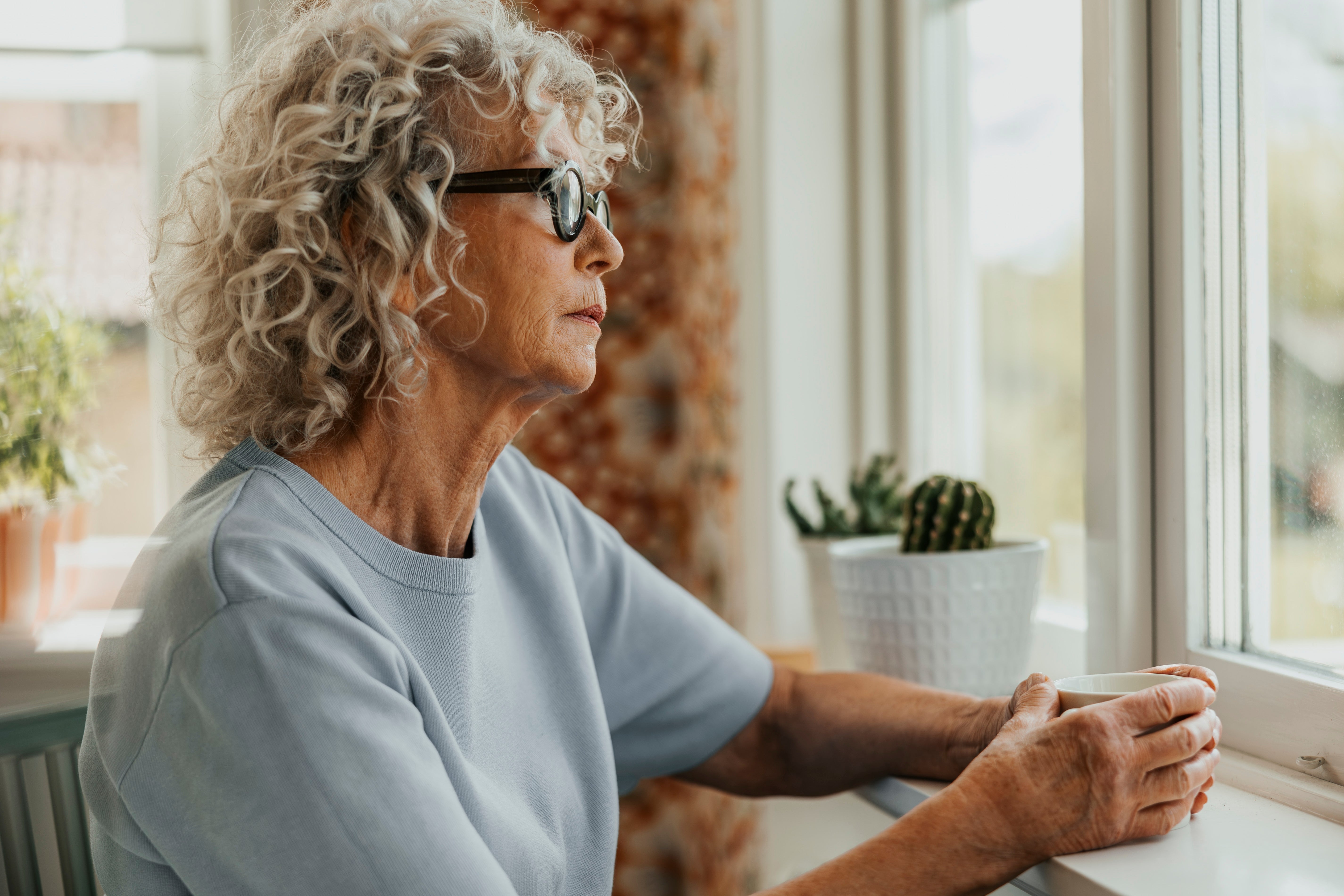 Elderly woman with curly hair gazes thoughtfully out a window, holding a mug, next to a potted cactus