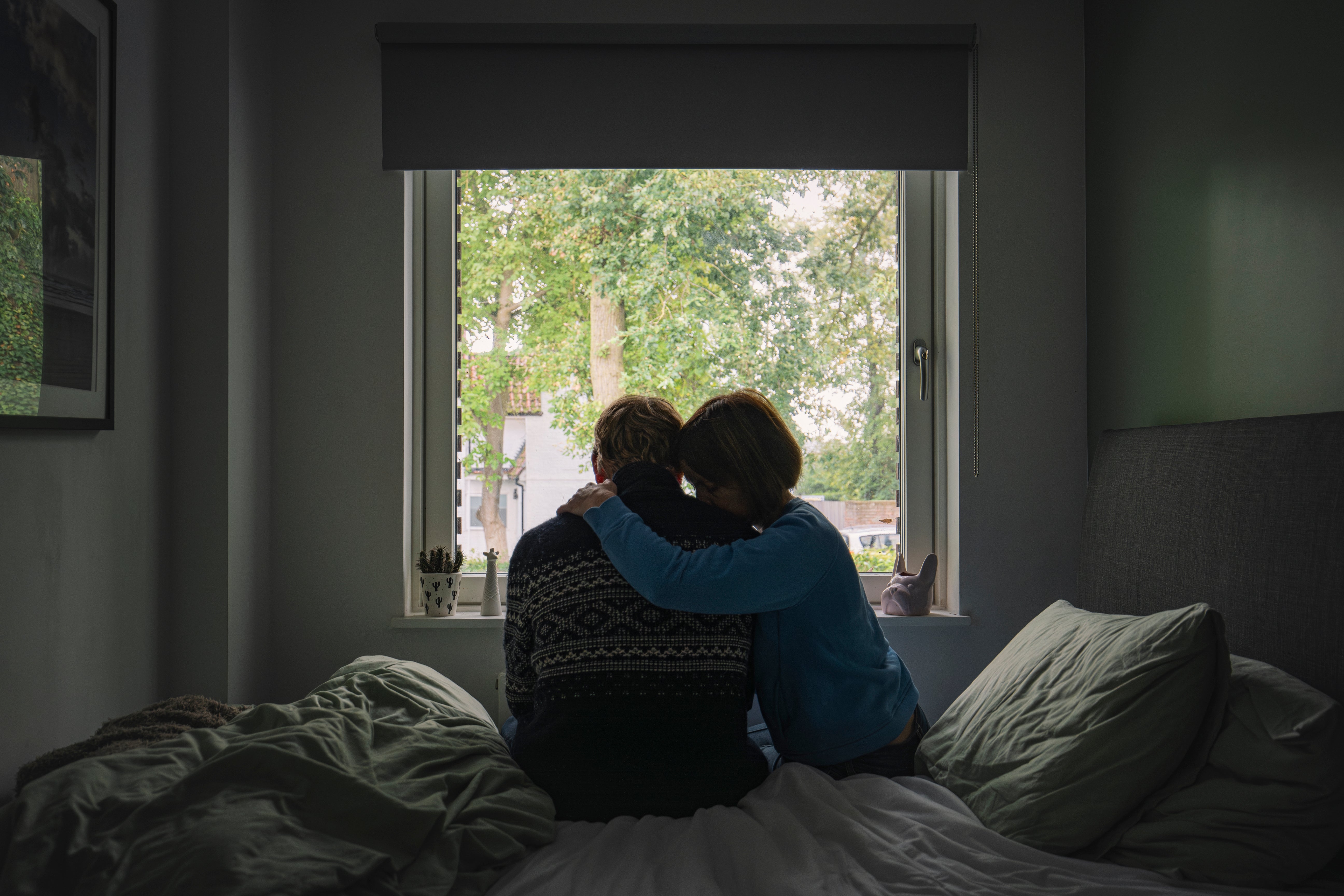 Two people sit on a bed, embracing, and look out a window at a tree-lined view, creating a comforting and contemplative scene