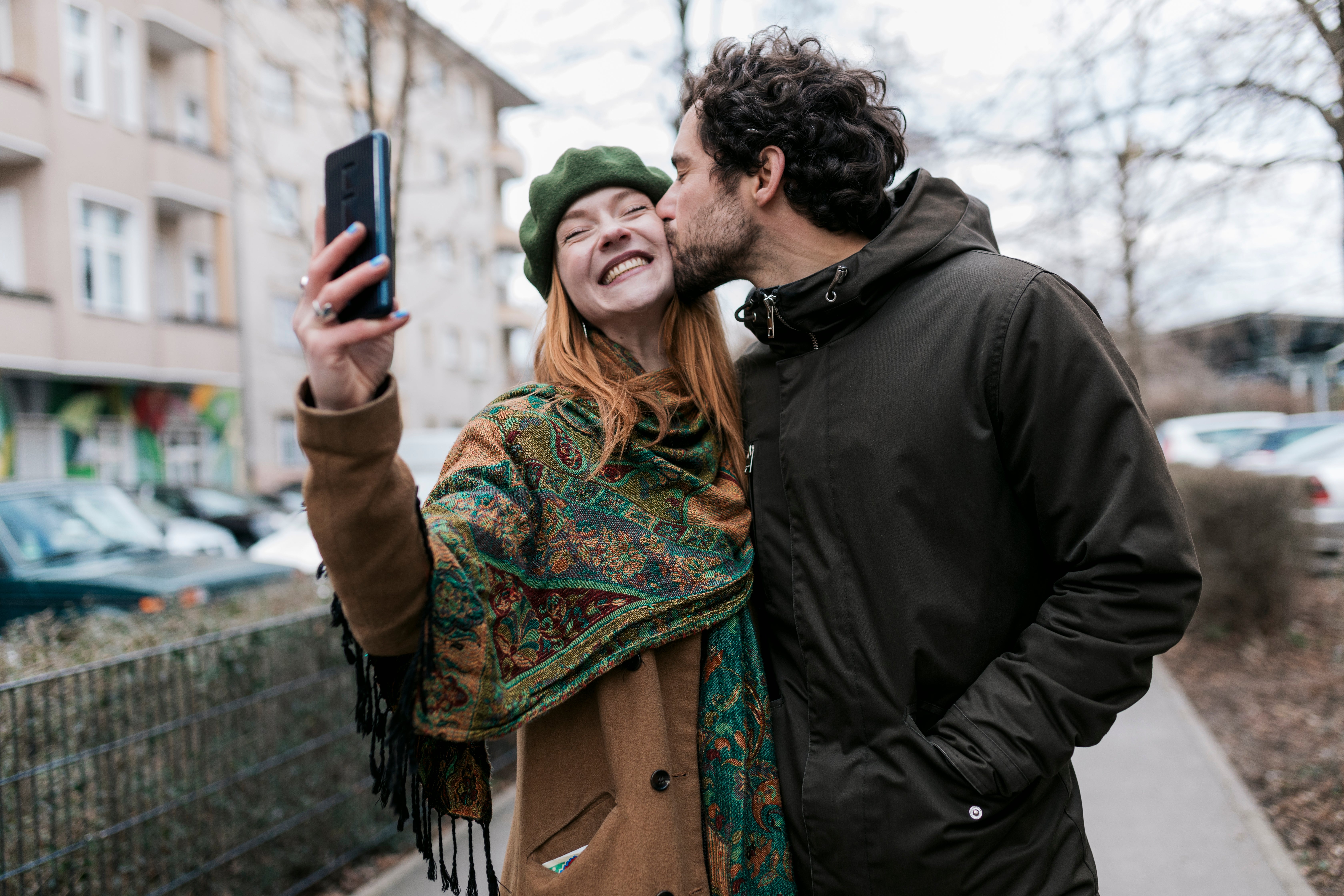 A couple taking a selfie outdoors, with one person kissing the other on the cheek. They look joyful and affectionate
